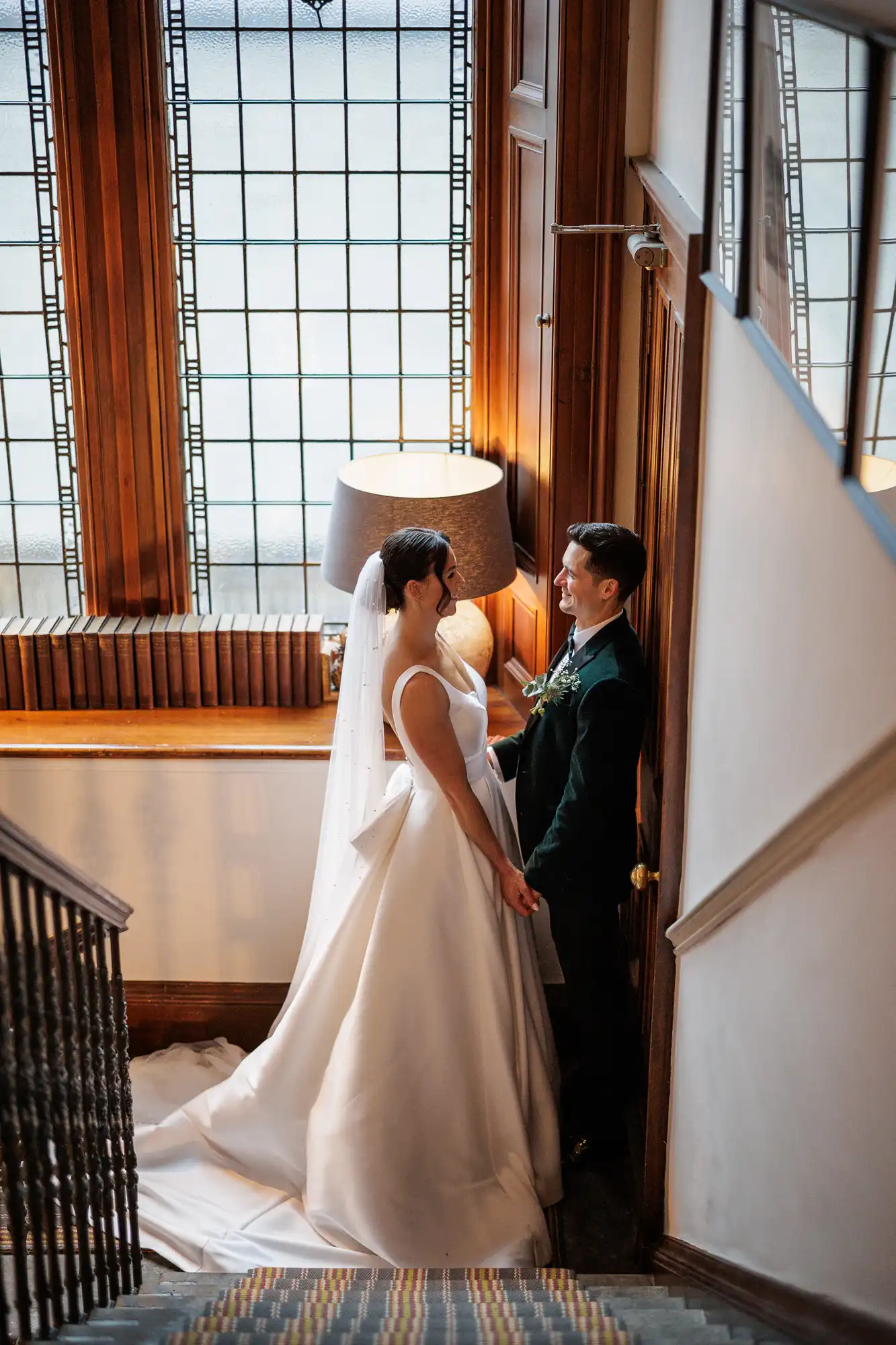 A bride in a white dress and veil, and groom in a dark suit, hold hands on a staircase, looking at each other.
