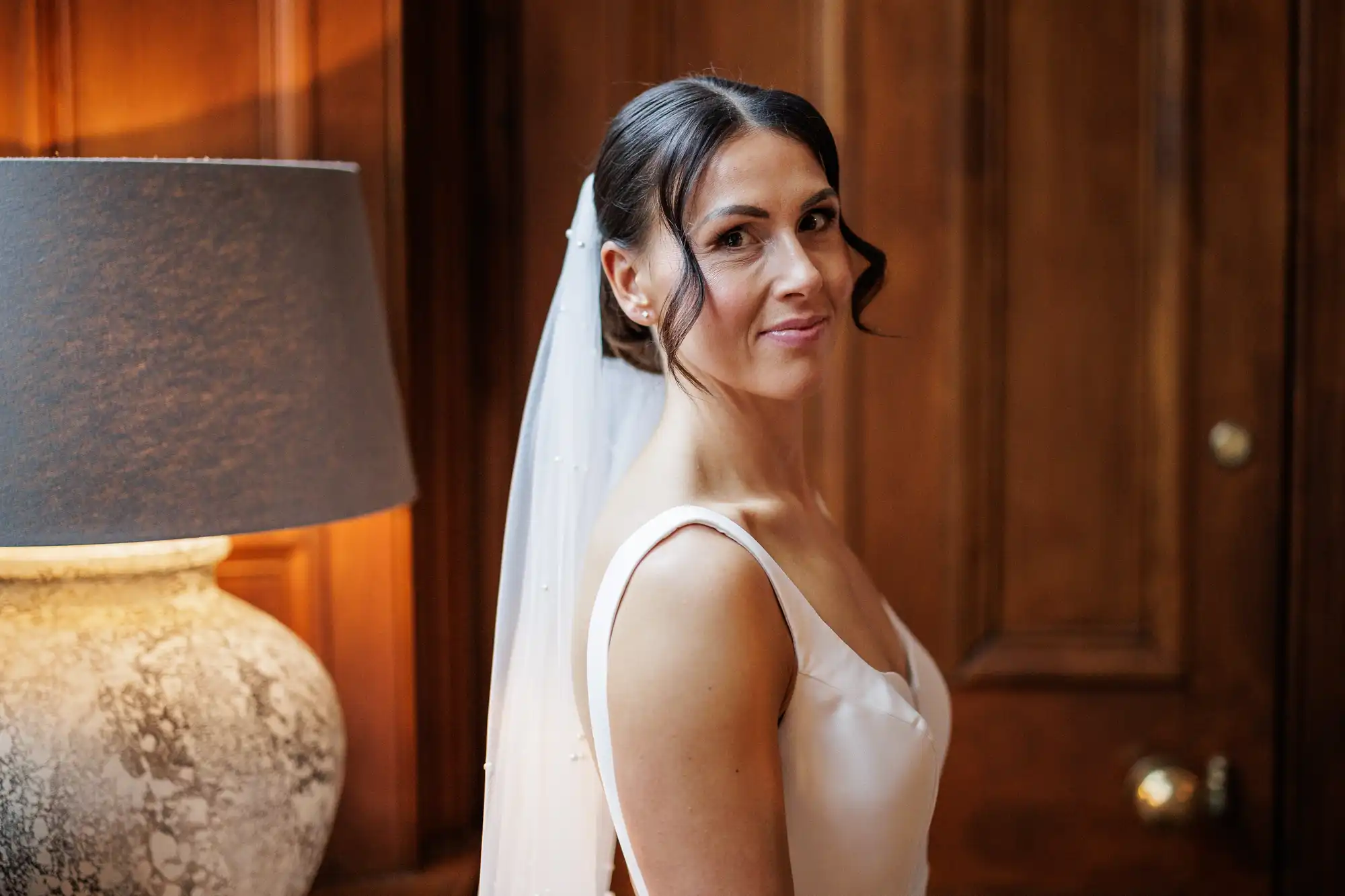 A bride with dark hair in an updo, wearing a white satin dress and a beaded veil, smiles over her shoulder. She is lit from the side, with a lamp visible to the left and a wooden door in the background.