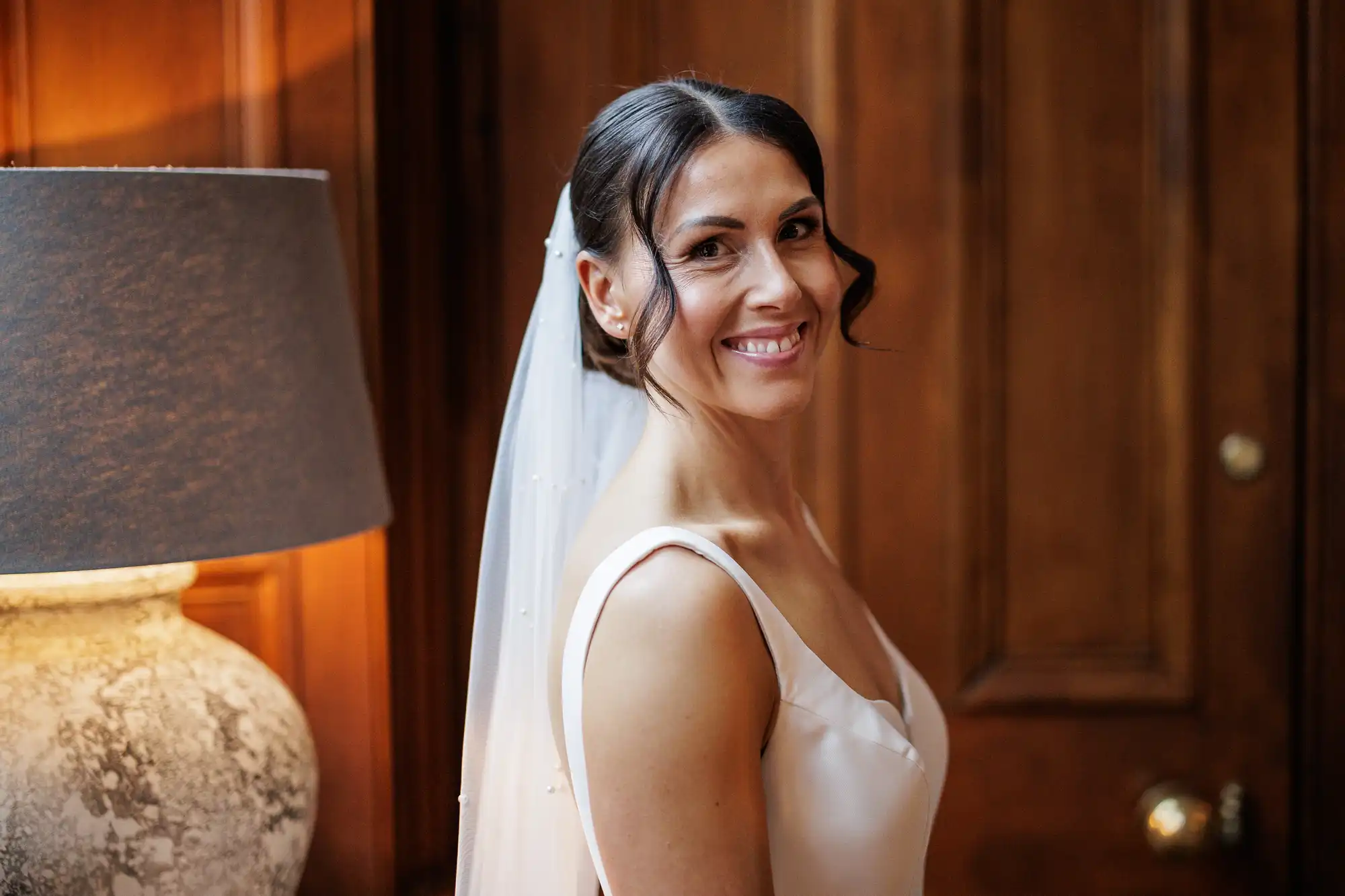Bride smiles, wearing a white dress and veil. She has dark hair styled up with some loose curls framing her face.