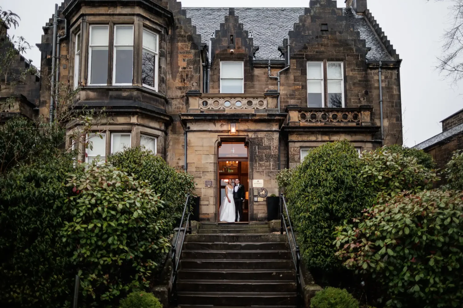 A bride in a white wedding dress and a groom in a tuxedo stand in the doorway of a stone building. Steps lead up to the entrance, flanked by lush green bushes.