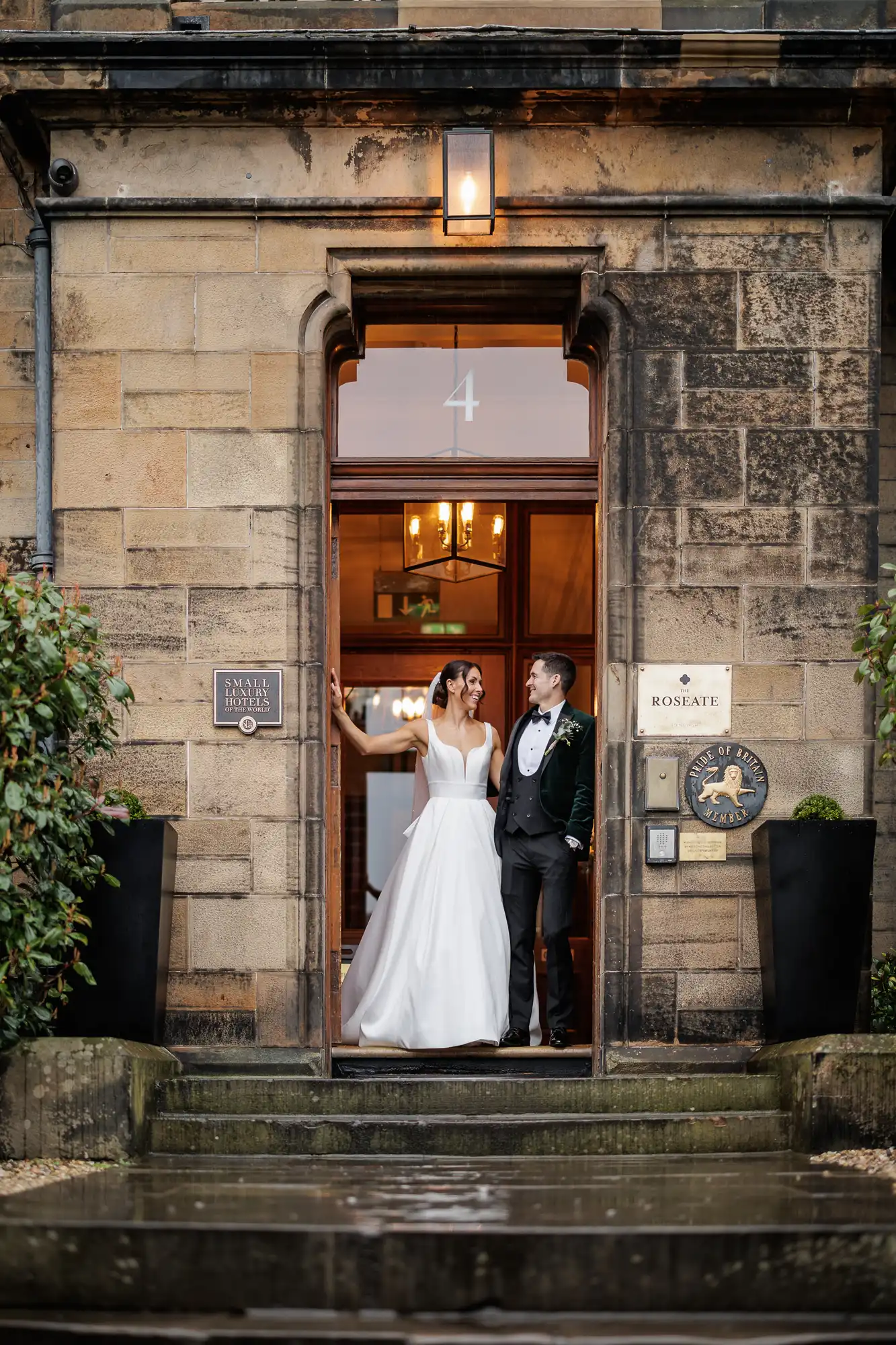 Newlyweds stand in a doorway of a stone building. The bride wears a white gown and veil. The groom wears a dark velvet tuxedo with a bowtie. Signs on the wall read "Small Luxury Hotels of the World" and "The Roseate Pride of Britain Member.