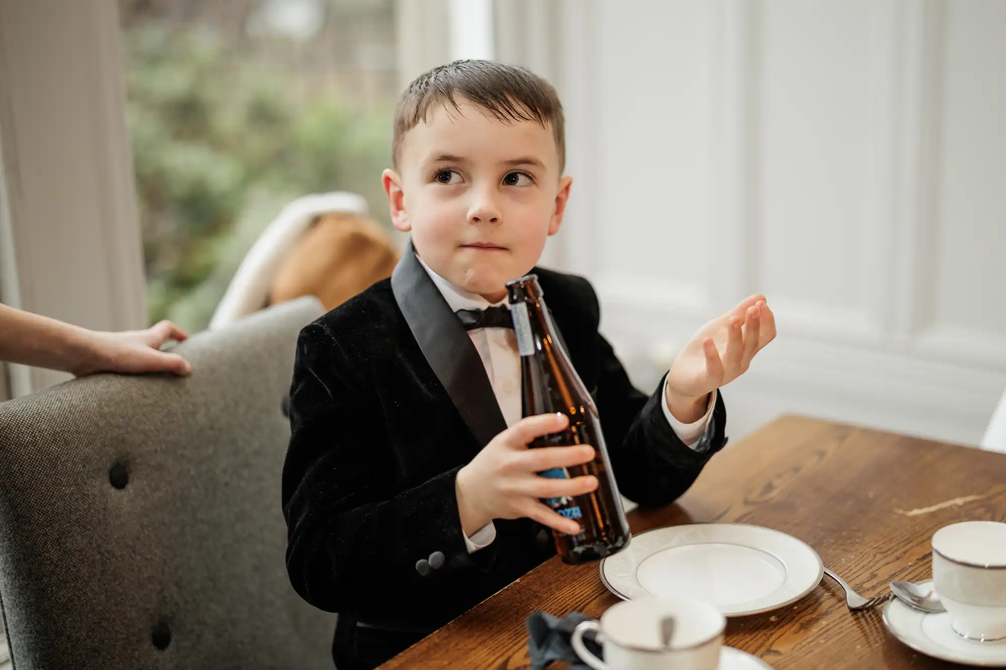 A young boy in a tuxedo holds a bottle, looking thoughtfully to the side. His left hand rests on the back of a chair, and his right hand gestures.