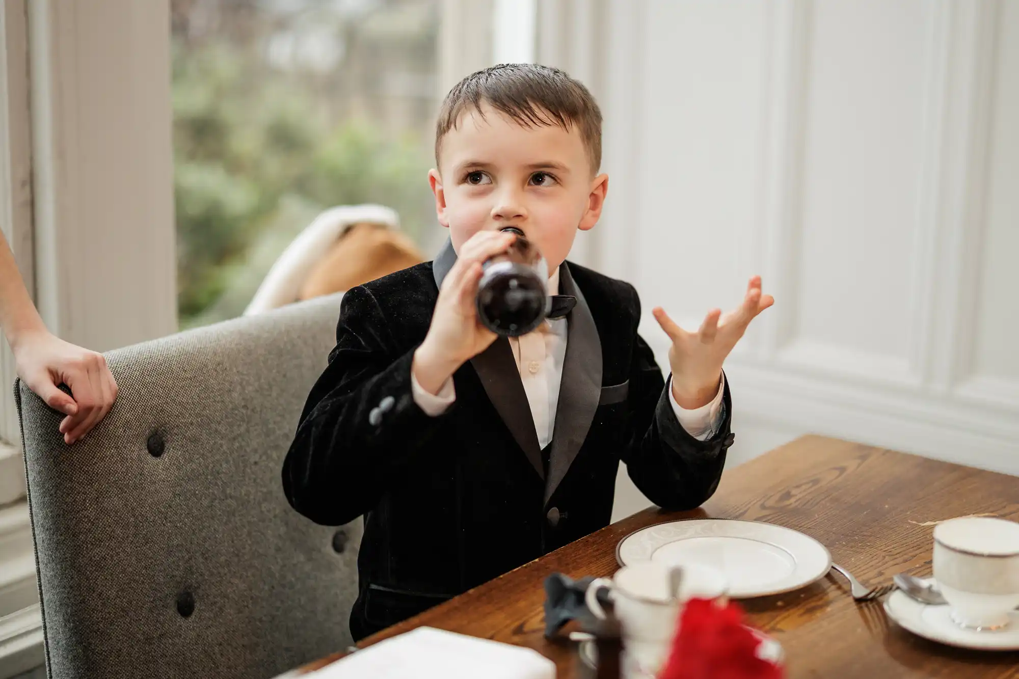 A young boy in a black velvet tuxedo and bow tie drinks from a dark bottle while gesturing with his free hand. He sits at a wooden table with a white plate and teacup.