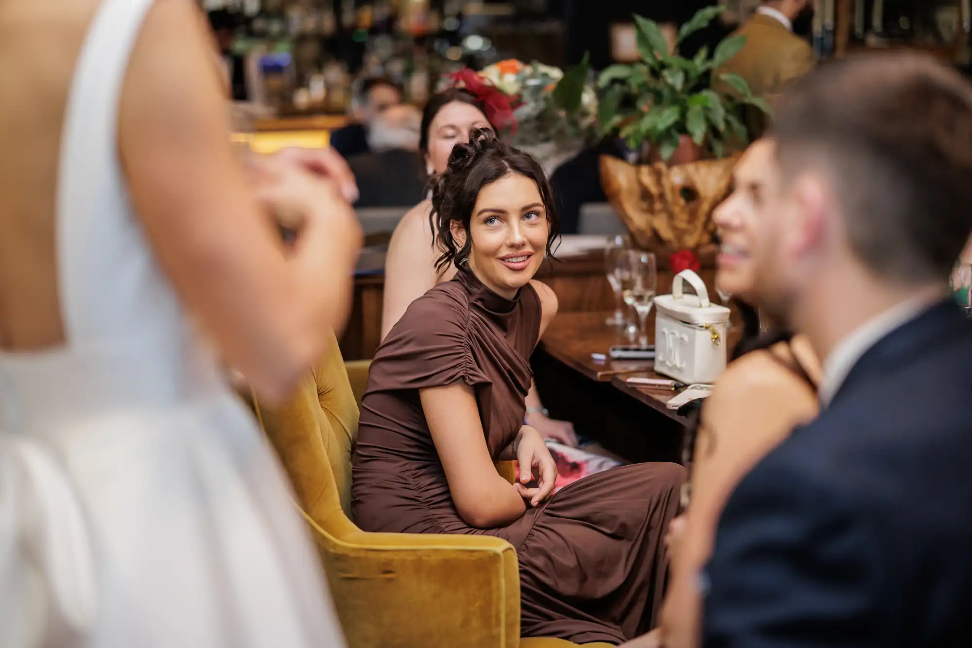 A woman in a brown dress sits in a yellow chair, smiling as she looks to her right. A person in a white dress is in the foreground on the left.