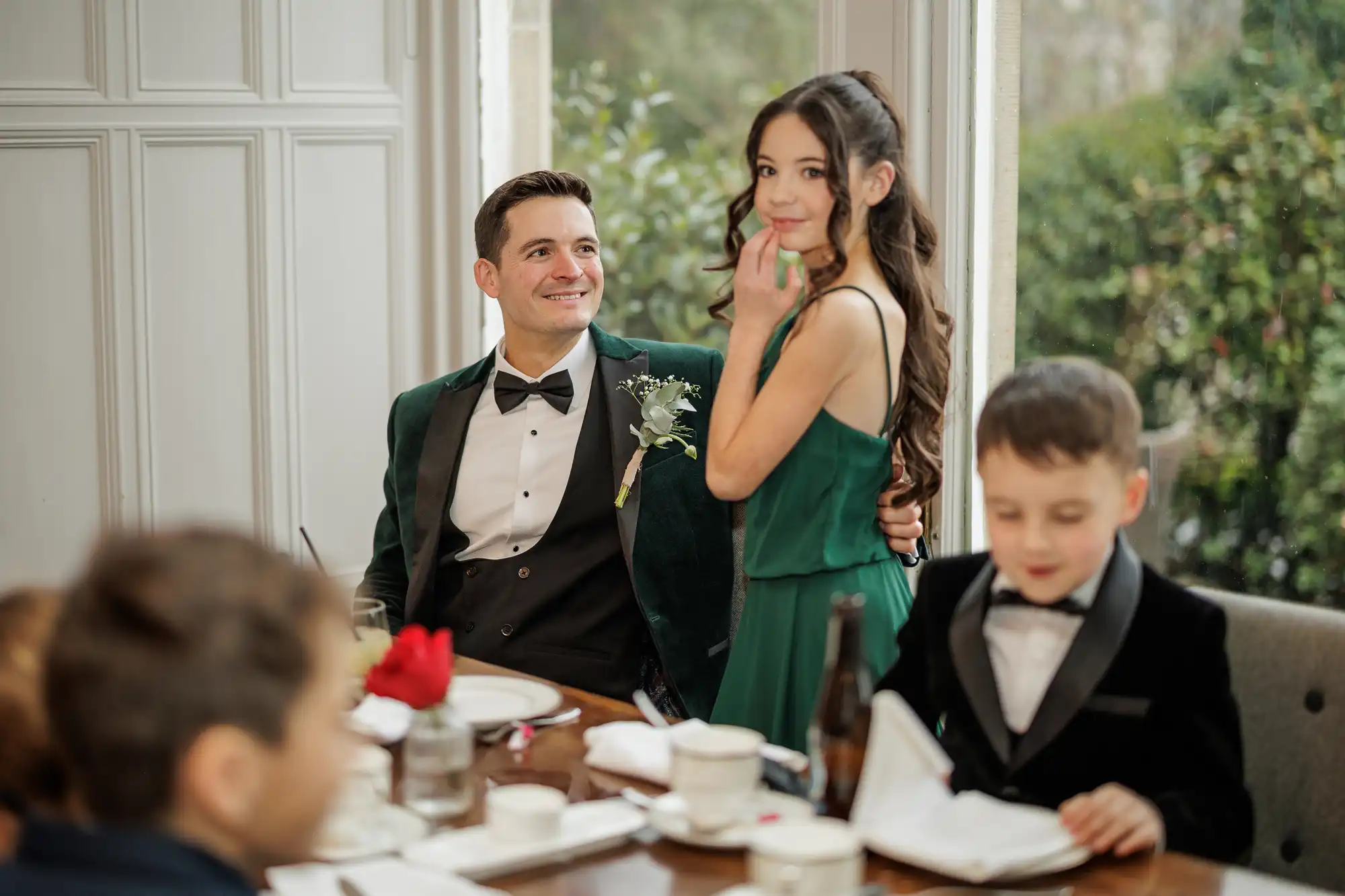 A man in a green velvet tuxedo smiles at a girl in a green dress. A boy in a black tuxedo sits at a table with a red rose and a drink.