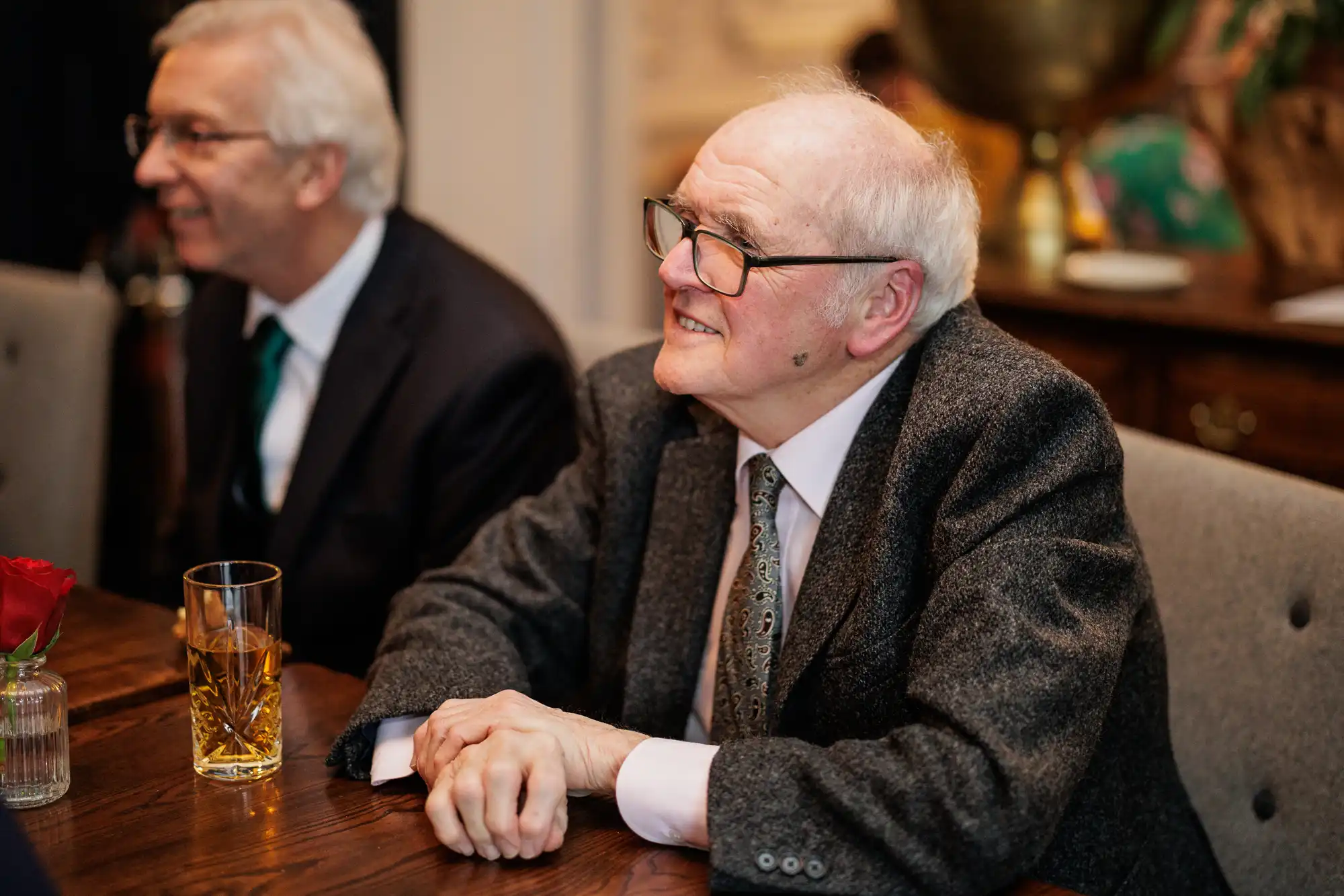 An elderly man with glasses and white hair smiles at a formal gathering. He wears a tweed jacket and patterned tie.