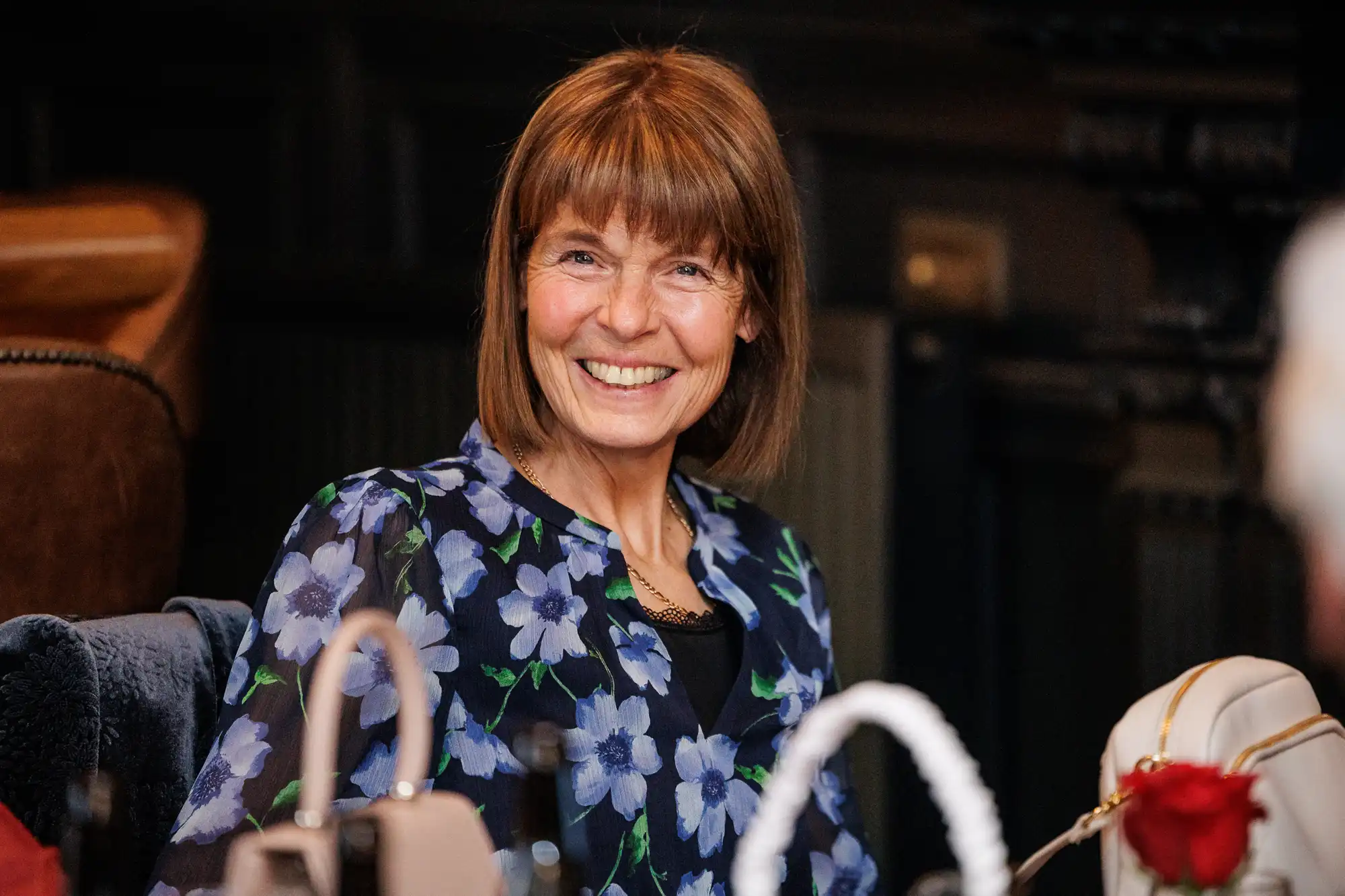 A smiling, middle-aged woman with chin-length brown hair and bangs, wearing a navy blue floral-print blouse and a gold chain necklace.
