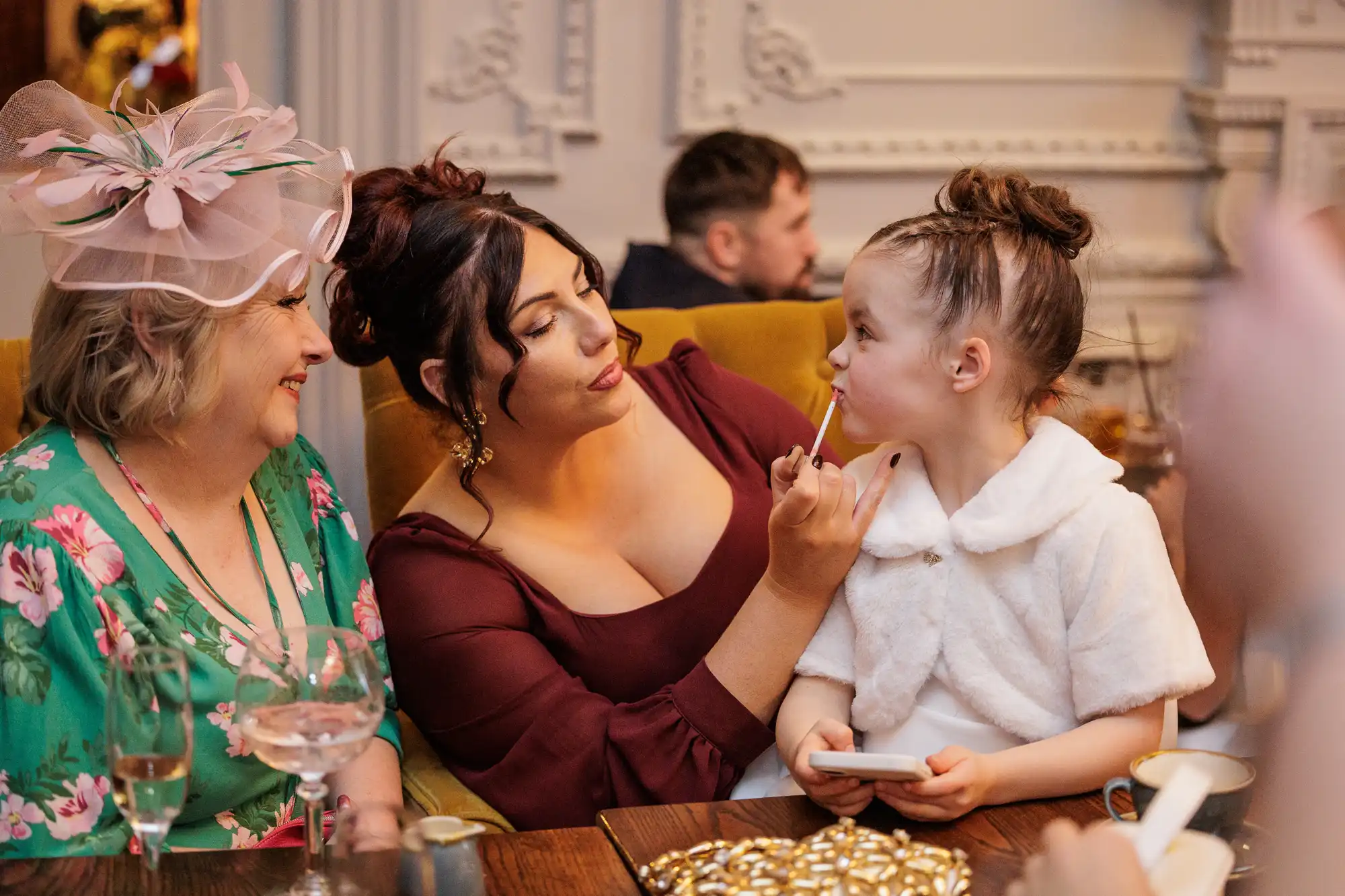 A woman in a burgundy dress applies lip gloss to a young girl in a white fur shrug. An older woman in a floral green dress and a large pink fascinator sits to the left.