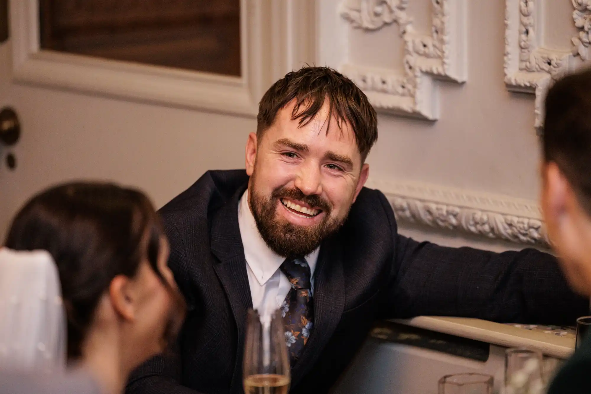 A bearded man in a suit and floral tie laughs joyfully at a formal event, leaning on a table with a champagne flute nearby.