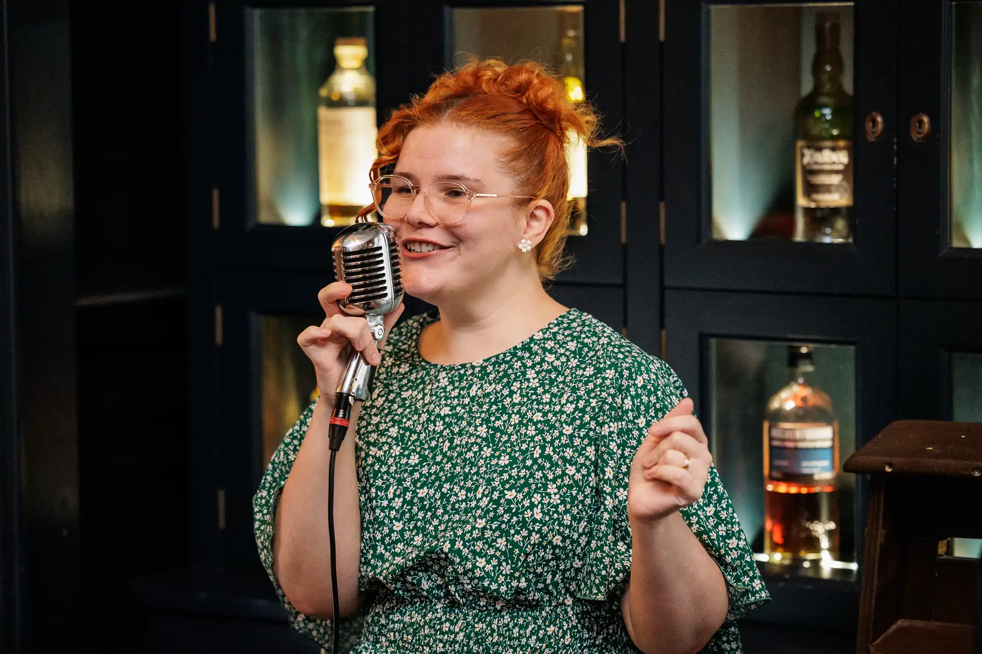 A woman with red hair and glasses holds a vintage microphone and speaks into it, wearing a green floral dress. Bottles of whiskey are displayed on shelves behind her.