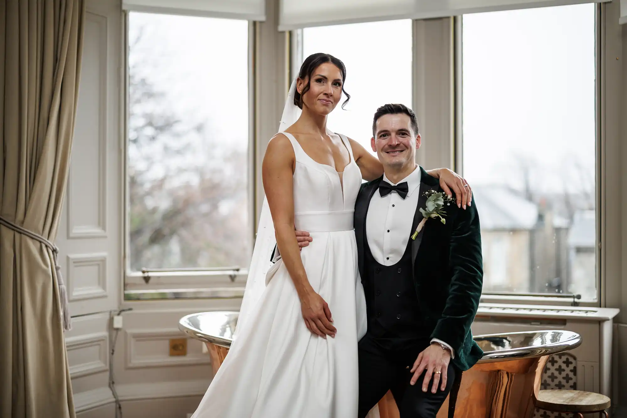 A bride in a white satin gown and a groom in a dark green velvet tuxedo smile at the camera, standing indoors with large windows behind them.