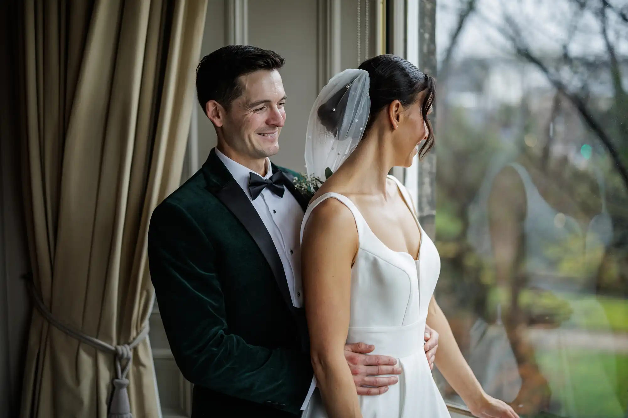 A bride in a white dress and a groom in a dark green velvet tuxedo embrace by a window. The groom looks at the bride, who is looking out the window.