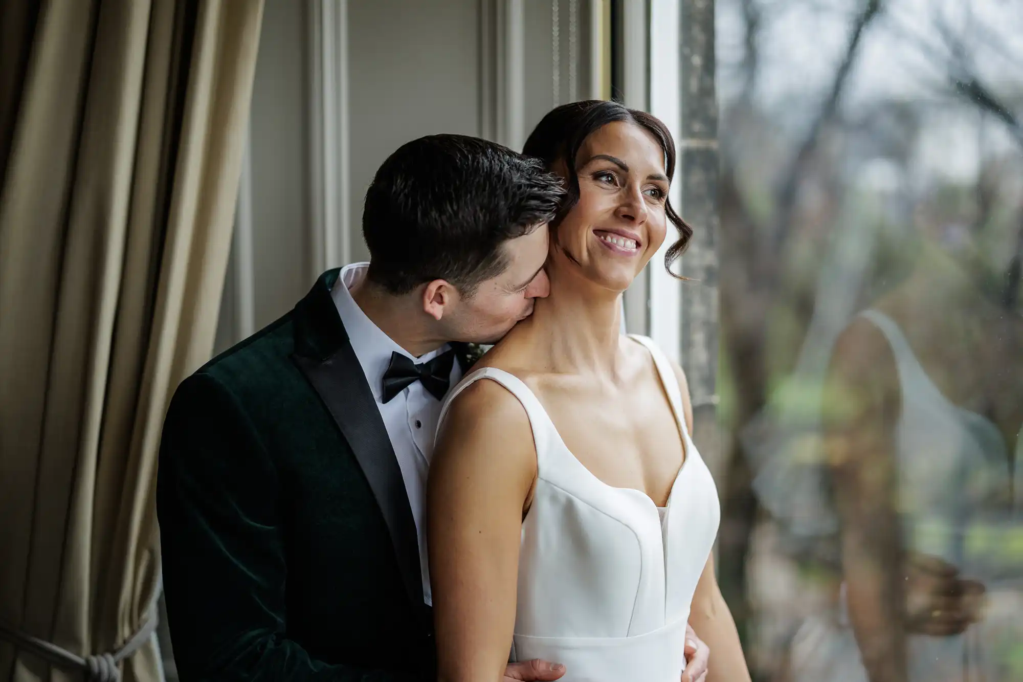Groom kisses bride's neck while she smiles looking out a window. She wears a white wedding dress and he wears a velvet green jacket and bow tie.