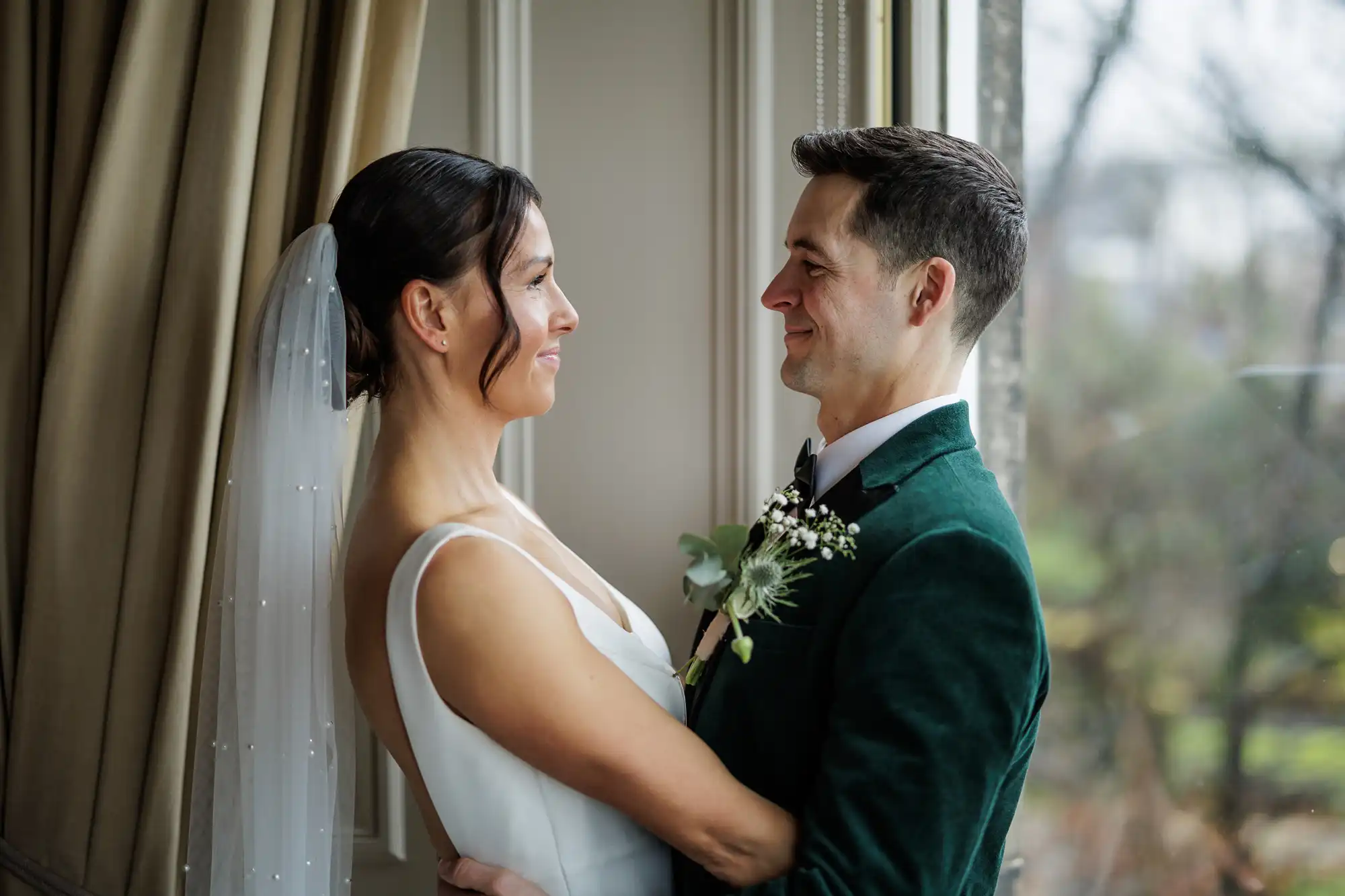 A bride in a pearl-embellished veil and white dress embraces a groom in a green velvet jacket, both looking at each other lovingly.