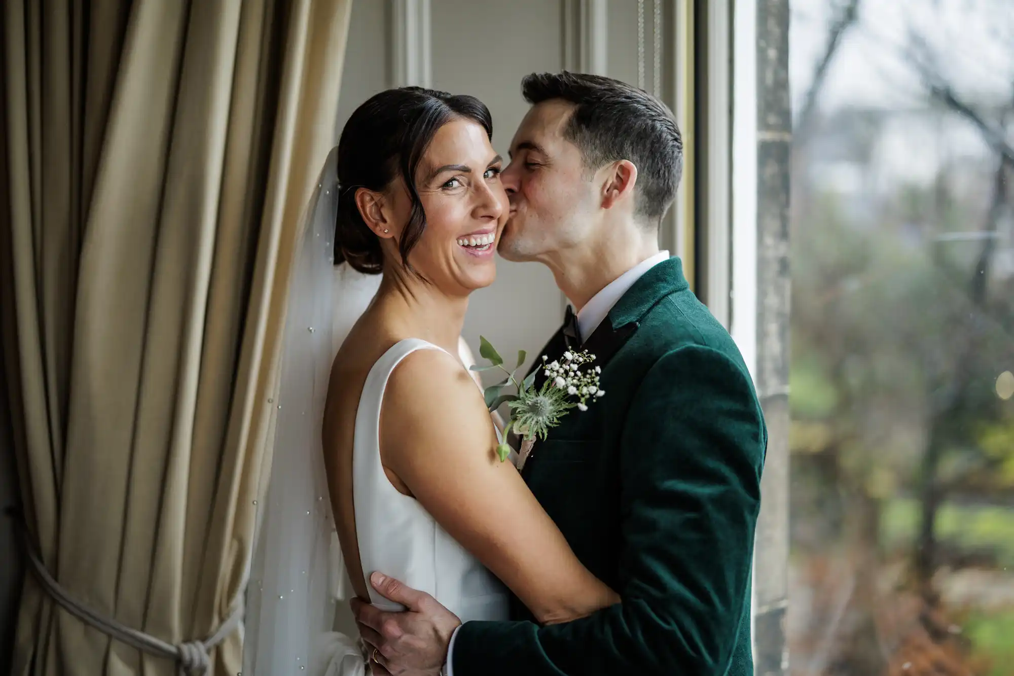A bride laughs as the groom kisses her cheek. She wears a white wedding dress and veil, he wears a dark green velvet jacket and bow tie.