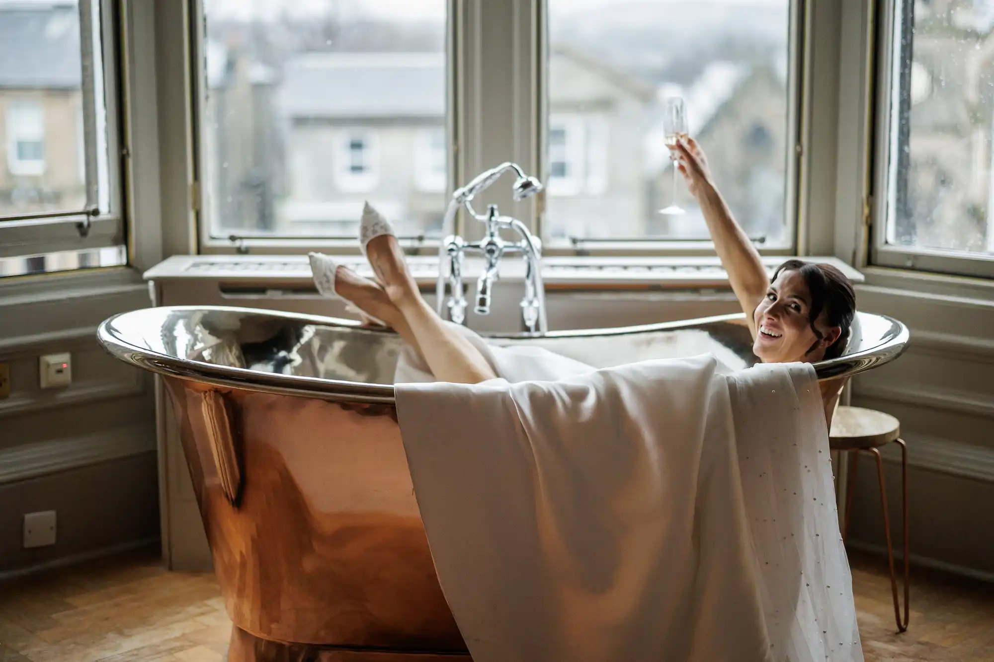 Bride relaxes in a copper bathtub holding a champagne flute. Her legs, clad in white lace heels, are propped up. A white veil with pearls drapes over the tub.