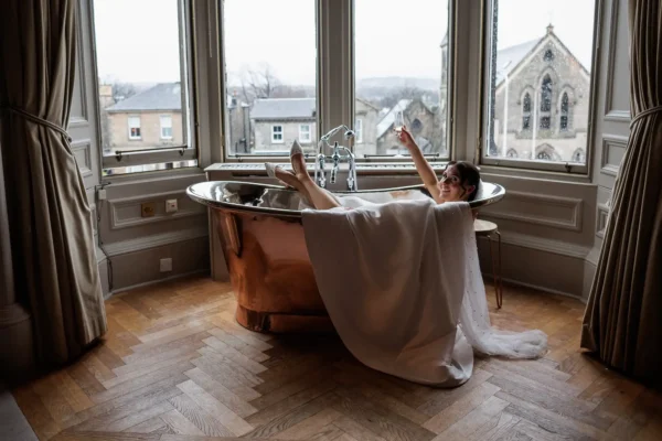 The Roseate Edinburgh wedding photographer image of a smiling bride in a bathtub, wearing heels and her wedding dress draped over her, holds up a glass of champagne. She is in a room with large windows overlooking a European town.