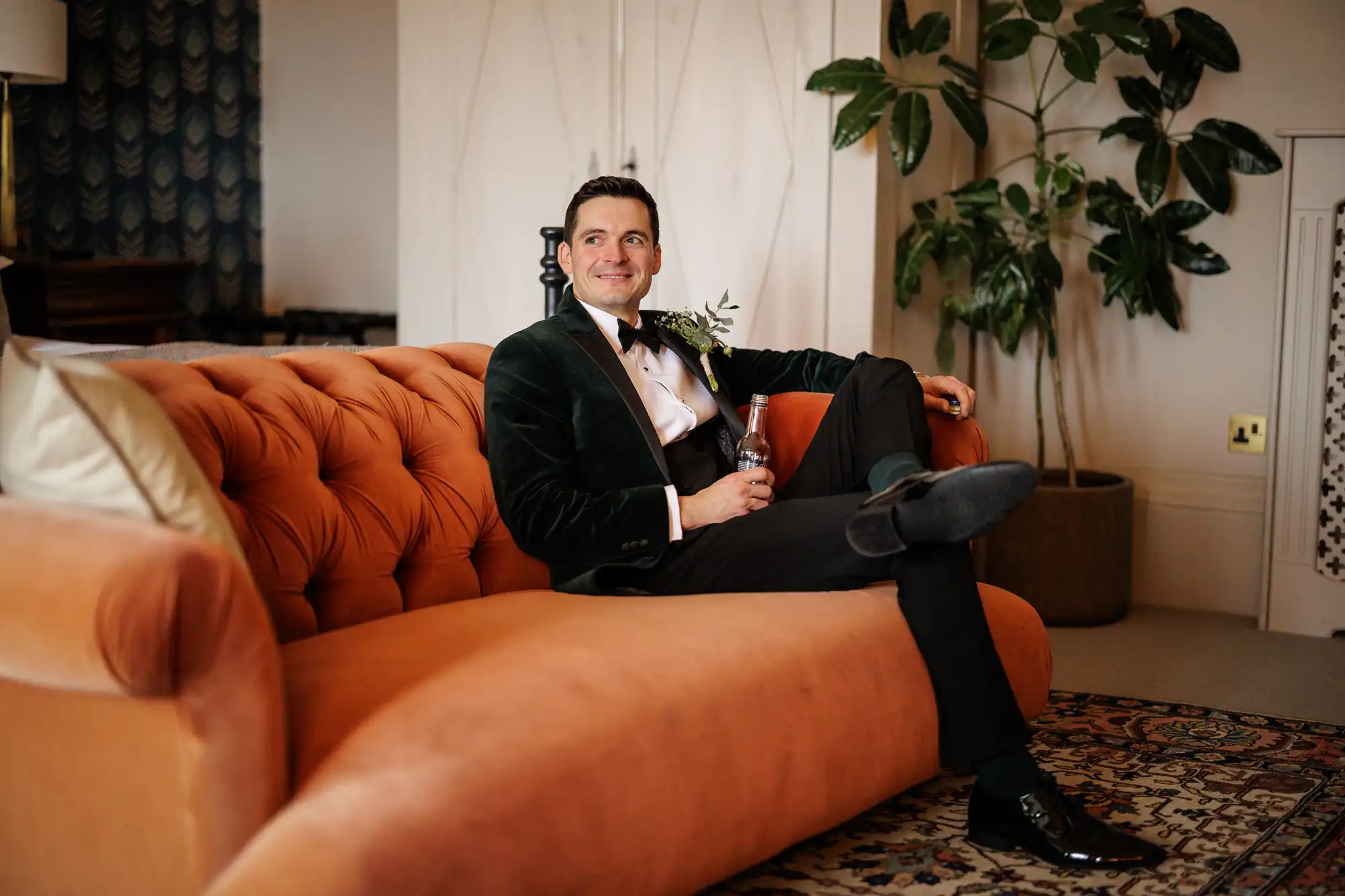 A groom in a velvet tuxedo jacket and bowtie relaxes on a plush orange sofa, holding a glass bottle. He smiles, with a boutonnière on his lapel.