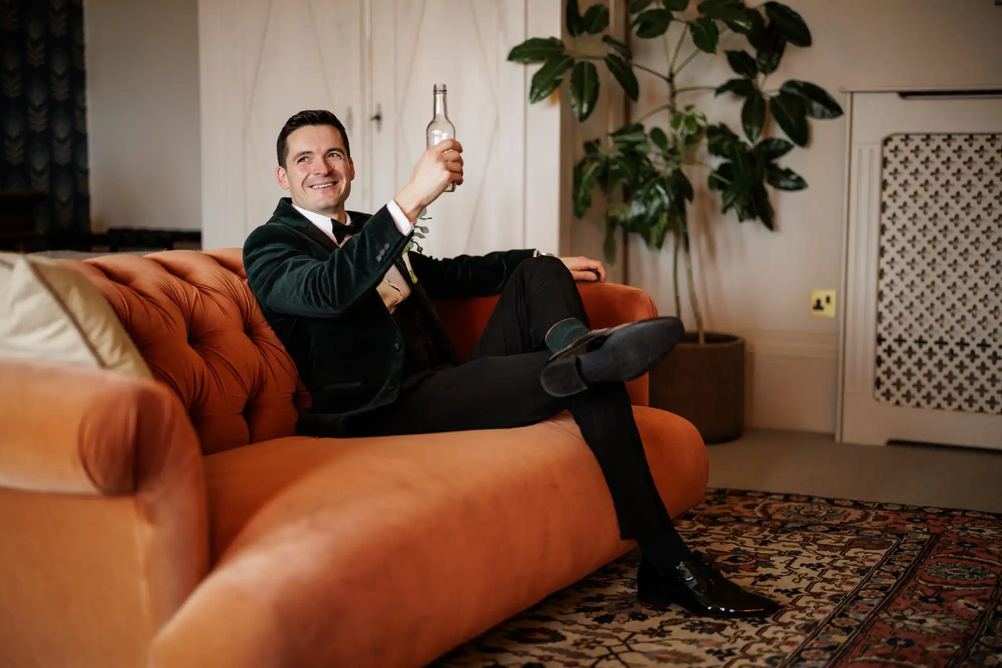 A groom in a velvet jacket, bow tie, and black pants sits on an orange couch, holding up a bottle. He smiles, looking off to the side.
