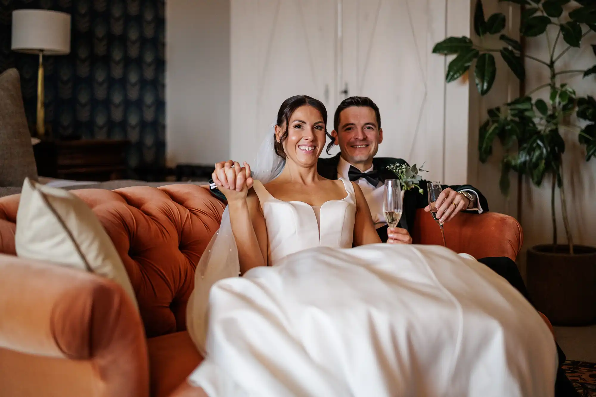 A smiling bride in a white wedding dress and a groom in a tuxedo sit close together on a rust-colored tufted couch, holding hands and champagne flutes.
