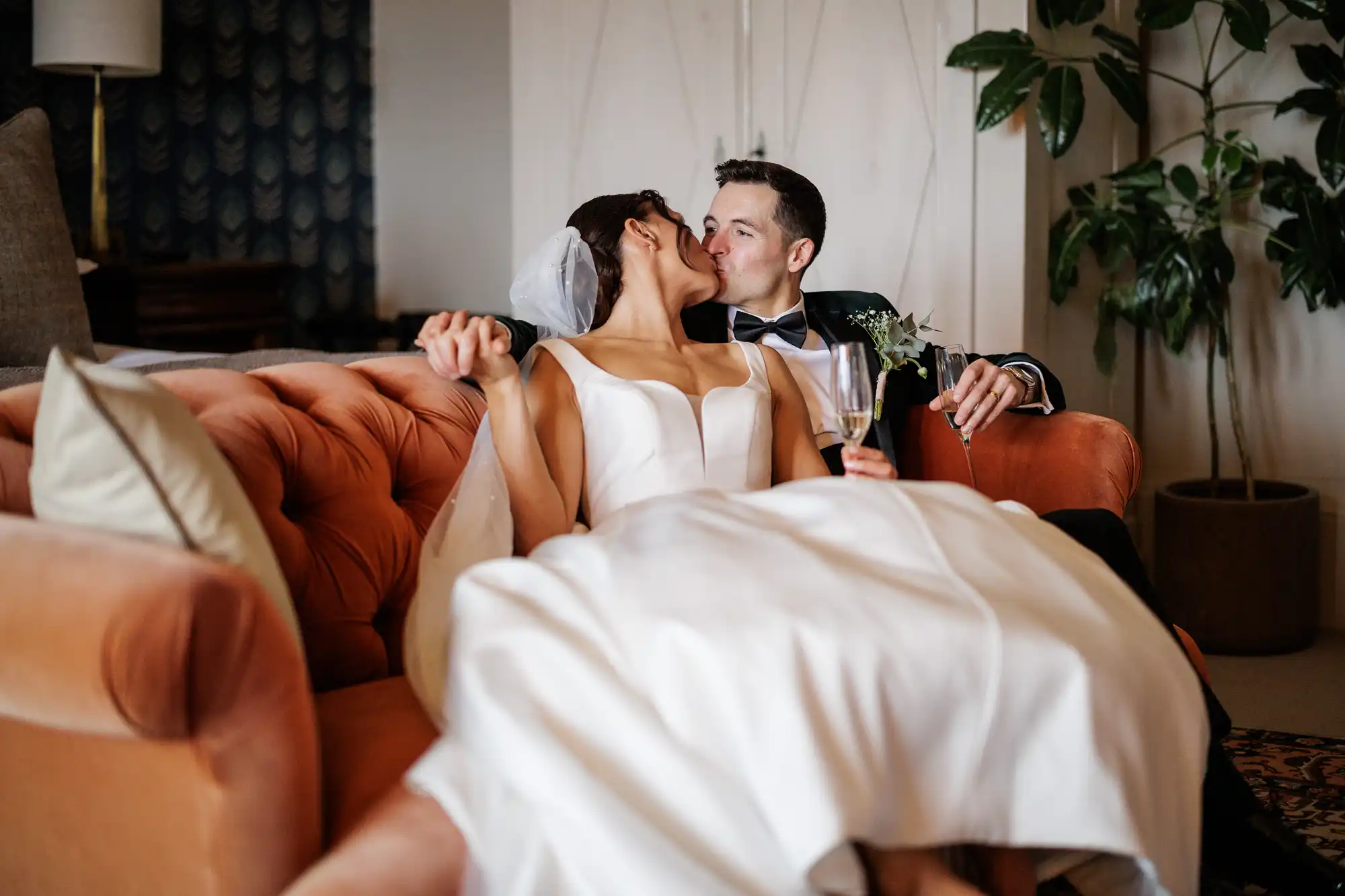 A bride and groom kiss on a plush orange couch. The bride wears a white dress and veil. The groom, in a tuxedo, holds two champagne flutes.