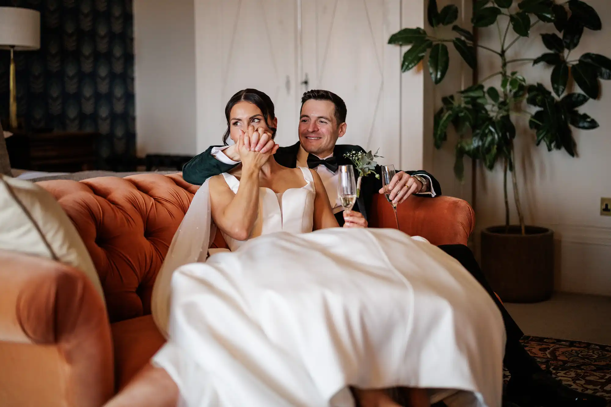 A groom in a dark green jacket and black bowtie smiles at his bride, who is wearing a white wedding dress and covering her mouth with her hands, as they sit on an orange couch holding champagne glasses.