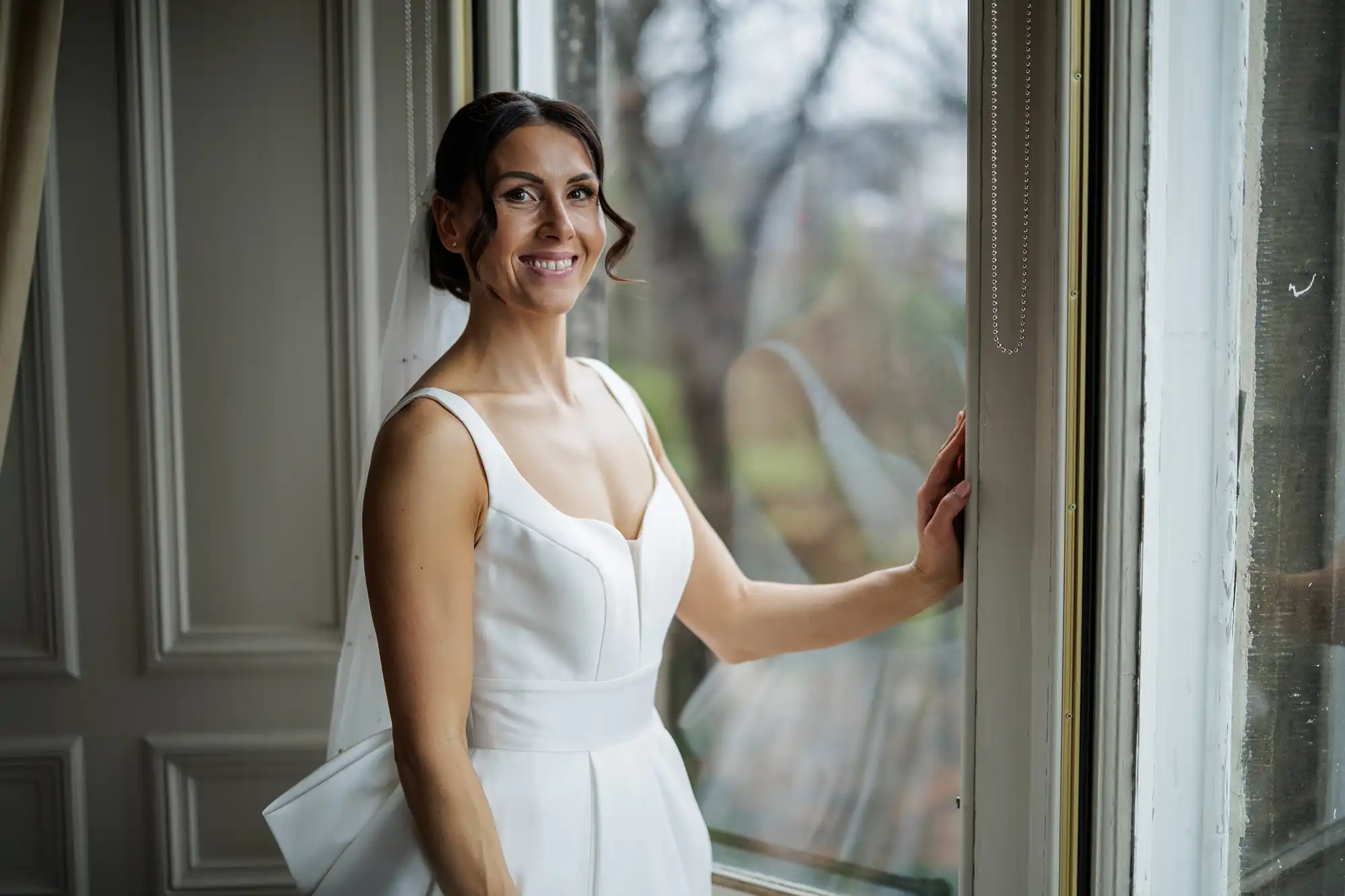 A bride in a white satin dress with a sweetheart neckline and veil smiles by a window.