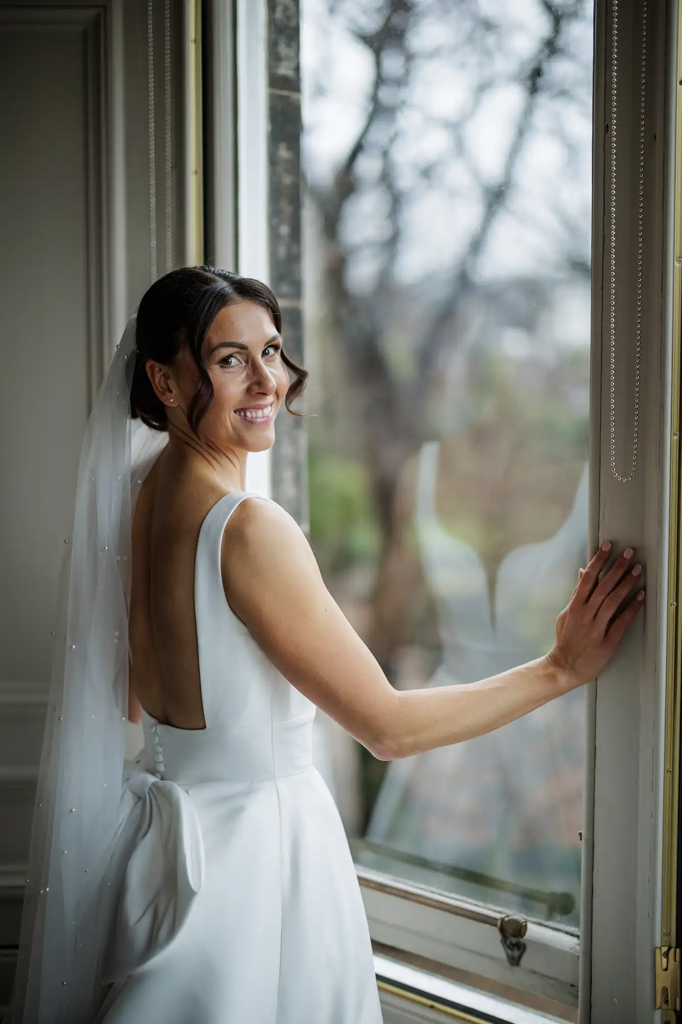 A smiling bride in a white, backless dress with a large bow looks out a window. Her veil is adorned with pearls.