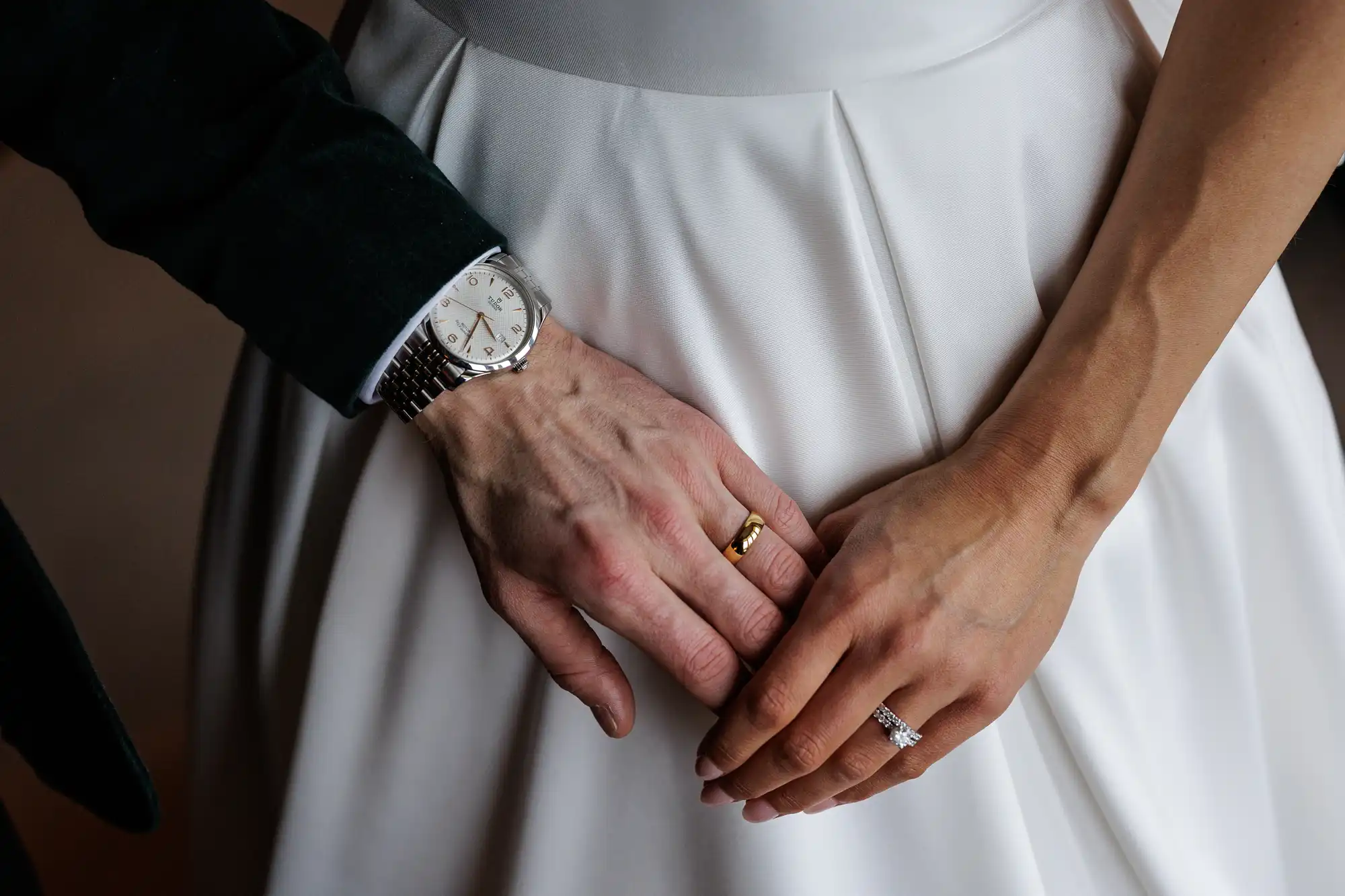 Close-up of a groom's hand wearing a watch and gold wedding band on his left hand, placed over a bride's hands, one wearing a diamond engagement ring, against her white wedding dress.