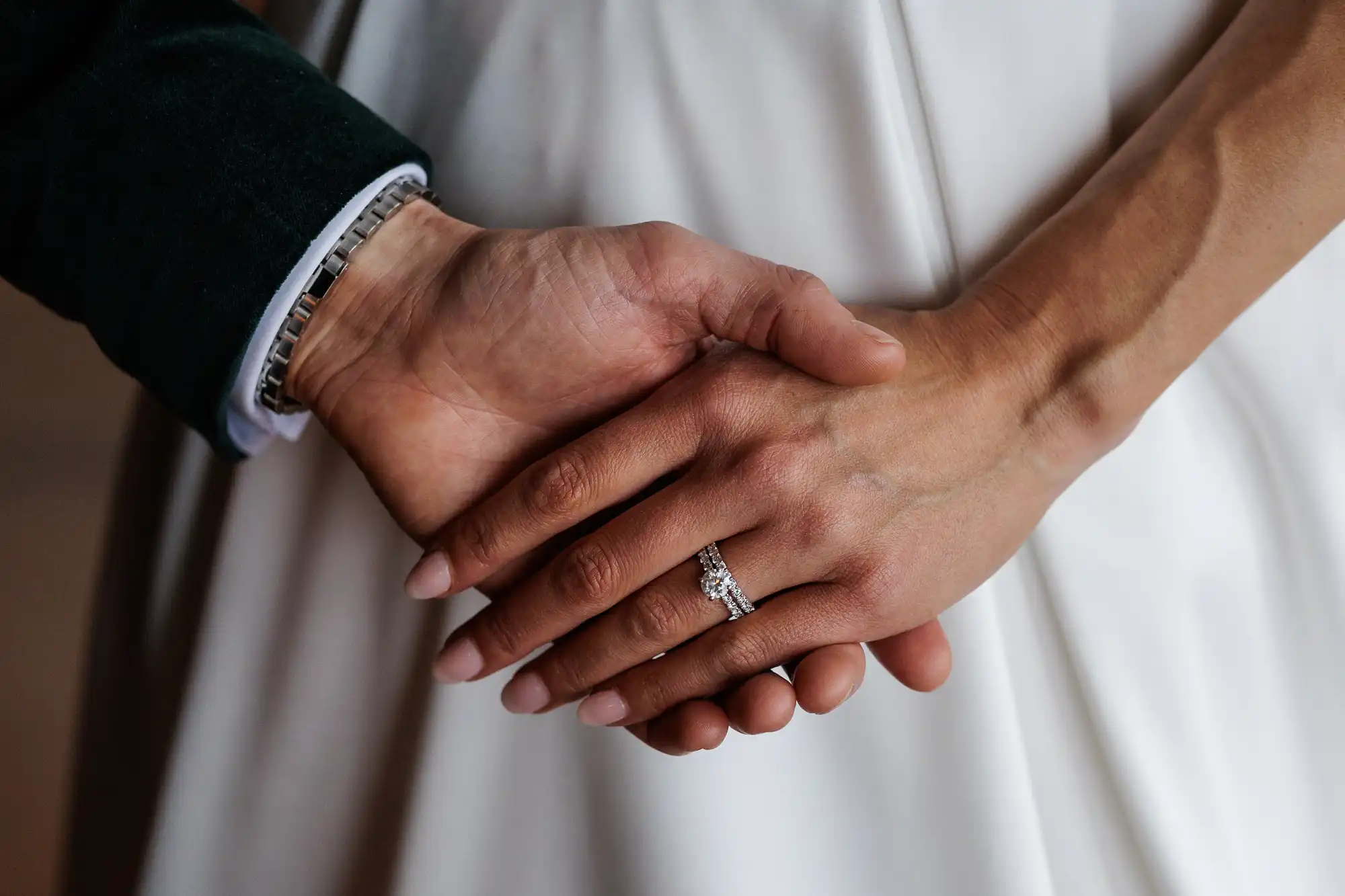 A groom in a dark green jacket and silver bracelet holds a bride's hands, adorned with an engagement ring, against her white wedding dress.