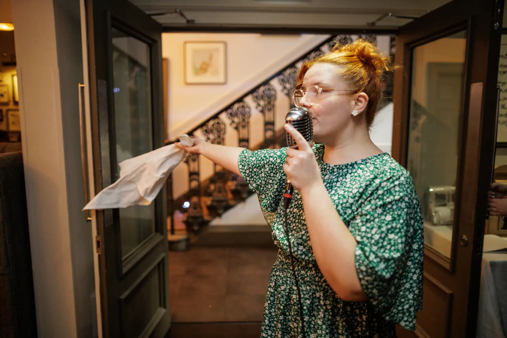 A woman with red hair in a bun, wearing glasses and a green floral dress, speaks into a vintage microphone while holding a white cloth. She stands in a doorway with a staircase in the background.