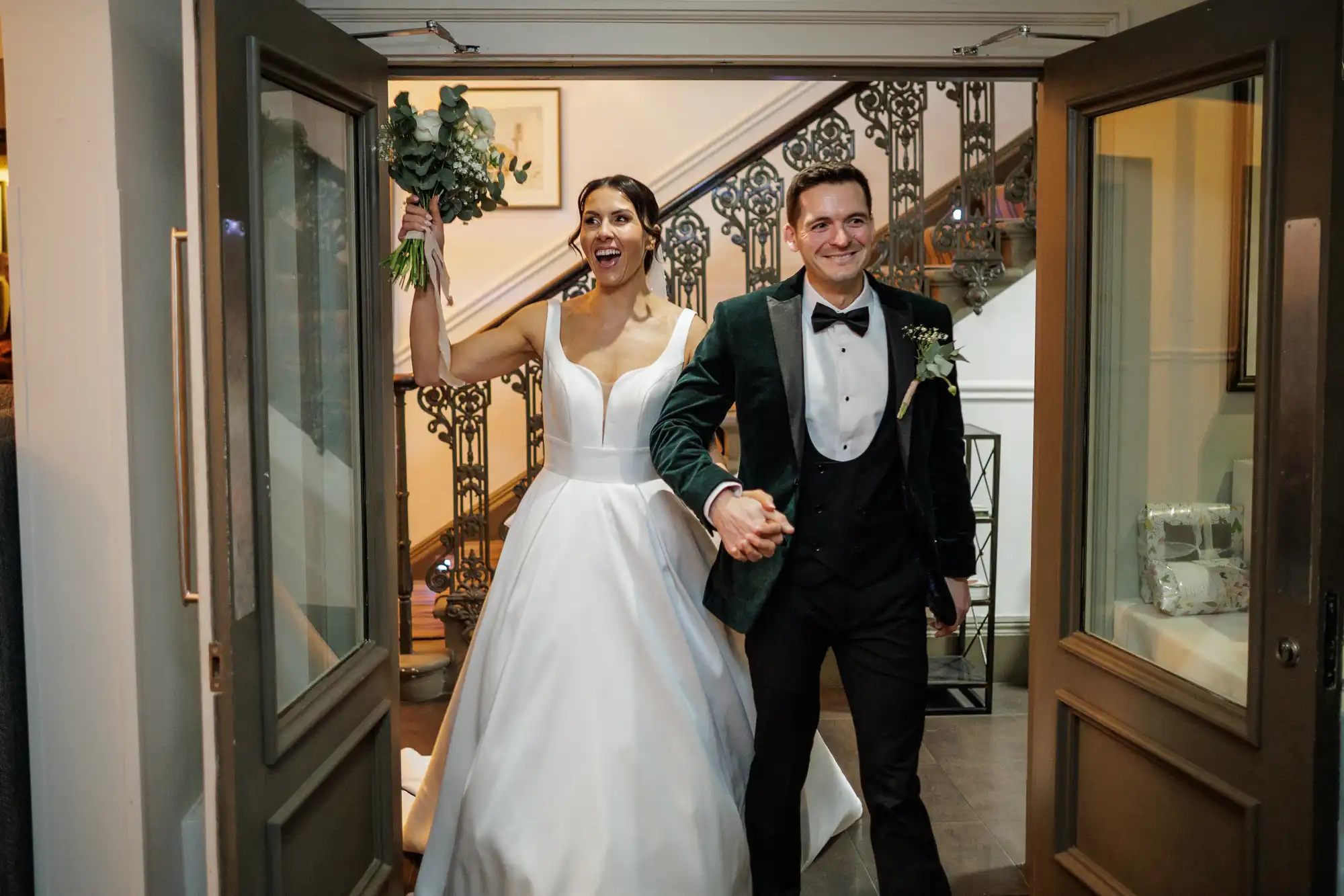 Bride and groom walk hand-in-hand through open doors. Bride smiles with her arm raised, holding a bouquet. Groom wears a green velvet jacket and bowtie.