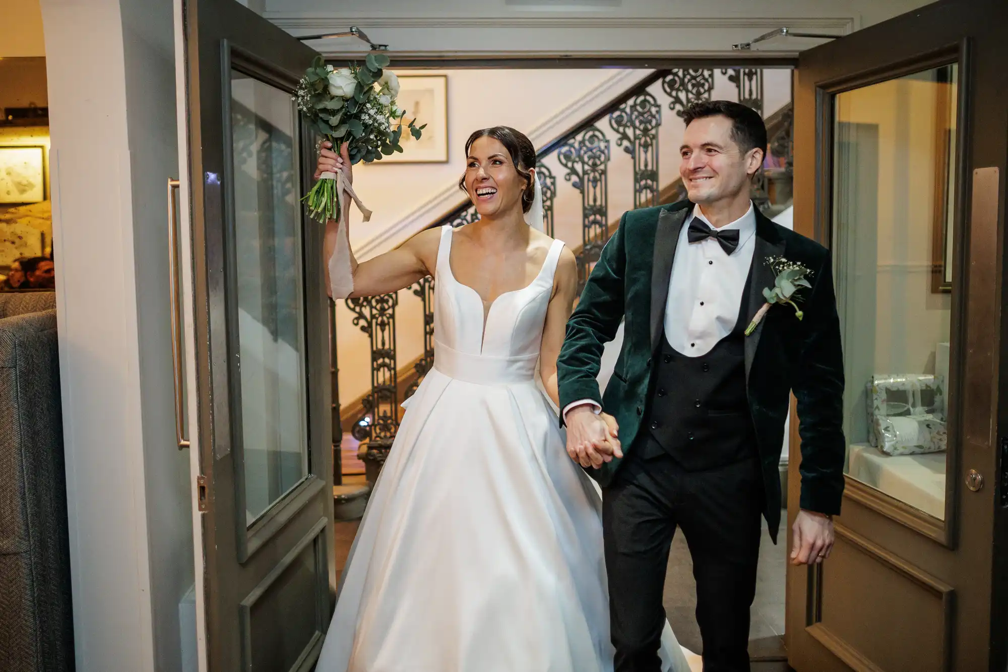 A beaming bride in a white gown and a groom in a velvet jacket hold hands as they enter through a doorway. The bride holds a bouquet of white roses.