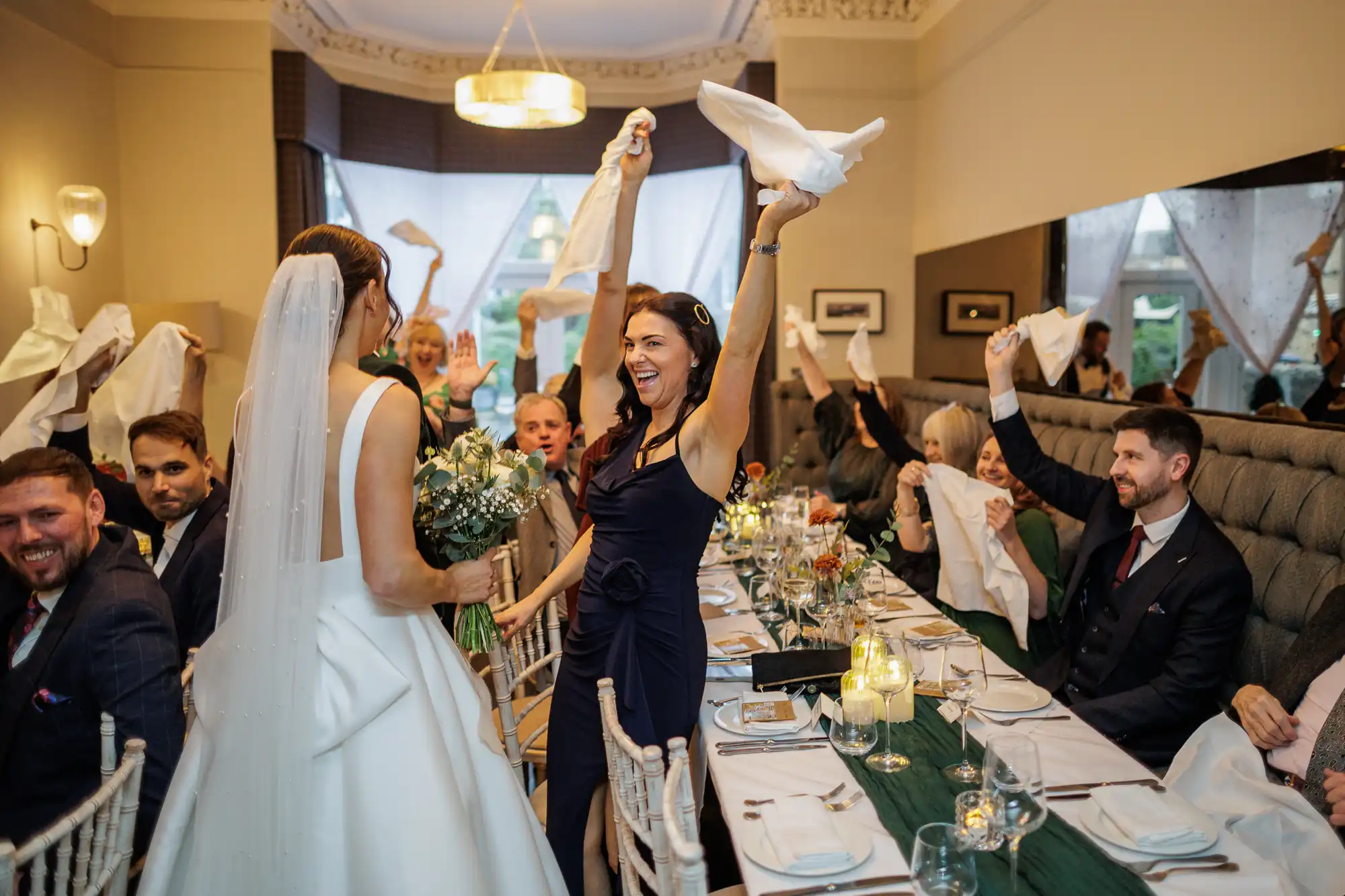 Wedding guests at a long table raise napkins in the air, celebrating. The bride in a white gown and veil stands in the foreground.