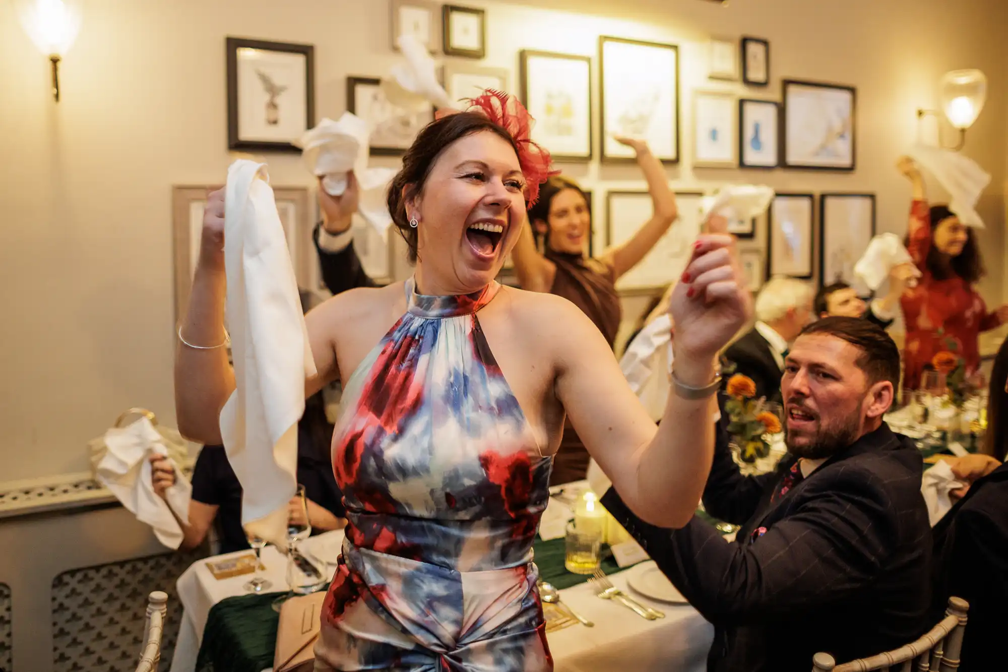 A woman in a colorful dress joyfully throws a white napkin in the air at a formal event. Other guests are also raising napkins.