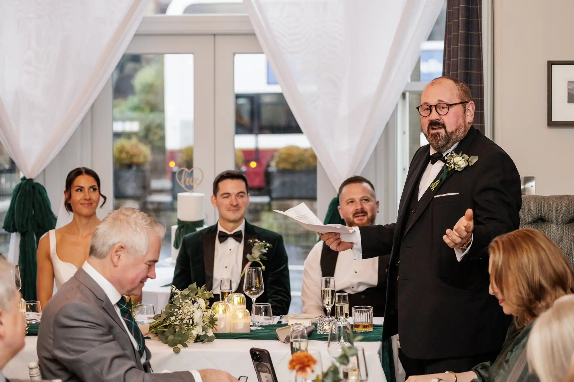 A man in a black suit with a bowtie and glasses gives a toast at a wedding reception. He stands before a wedding party, including a bride in a white gown and groom in a velvet jacket.