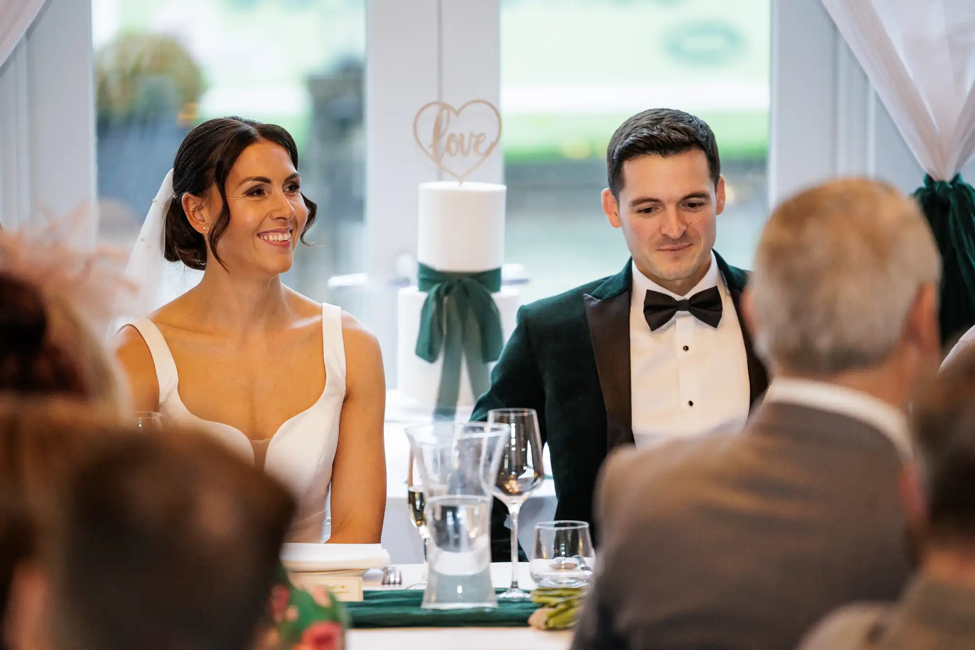 A bride in a white dress and groom in a velvet tuxedo smile at their wedding reception, seated with guests.