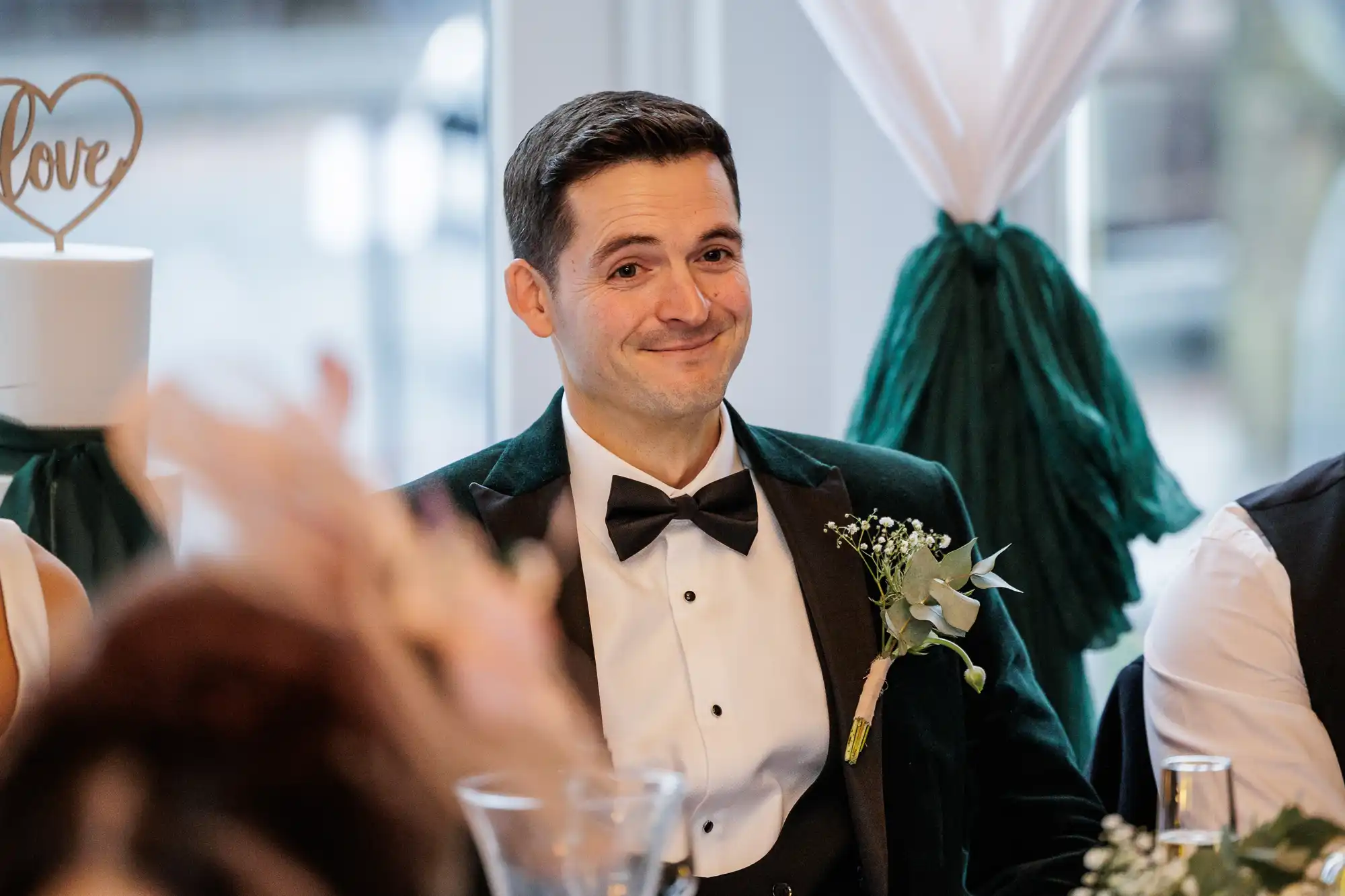 A groom smiles, wearing a dark green velvet jacket, white shirt, and black bowtie. A boutonnière with eucalyptus and baby's breath is pinned to his lapel.