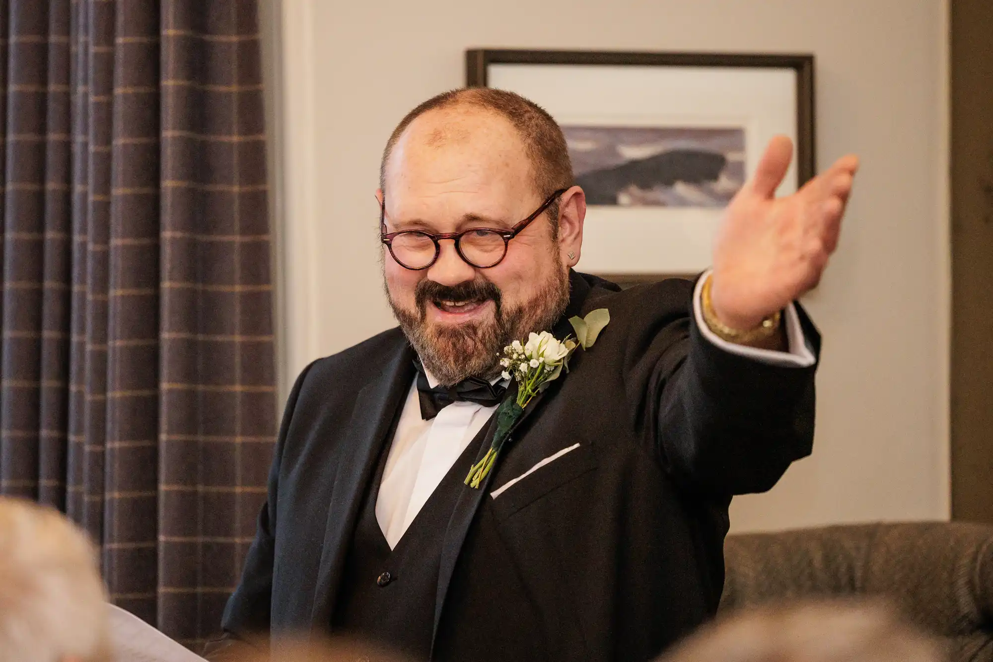 A smiling, bearded man in a tuxedo and bow tie speaks animatedly with his right hand raised, wearing glasses and a boutonniere.