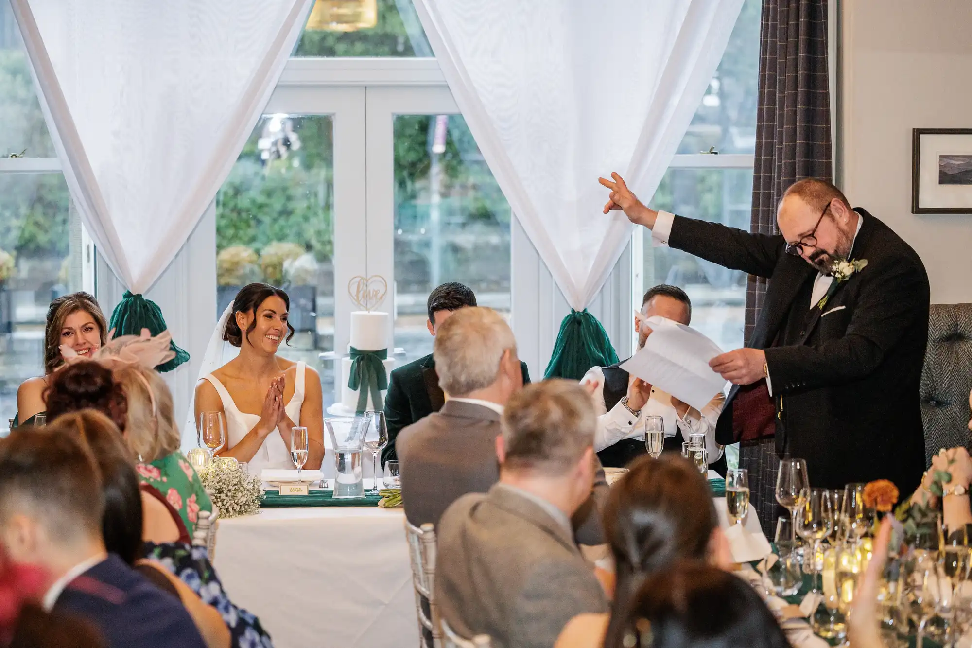 A wedding reception speech. A man in a suit gives a speech while holding papers. The bride smiles and clasps her hands. Bridesmaids and guests sit at tables decorated with flowers and wine glasses.