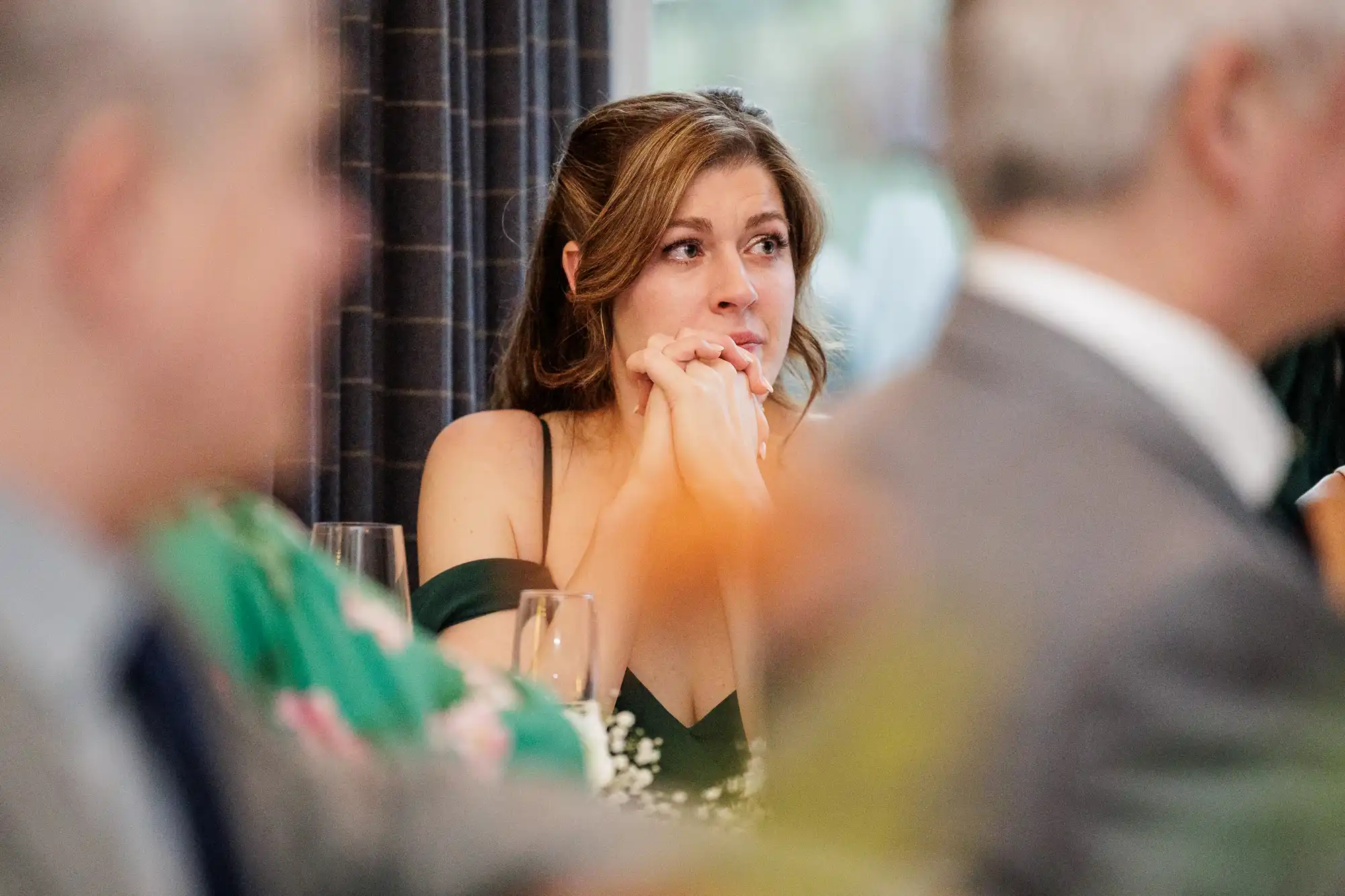 A young woman with brown hair and a green dress looks away with her hands clasped near her chin.