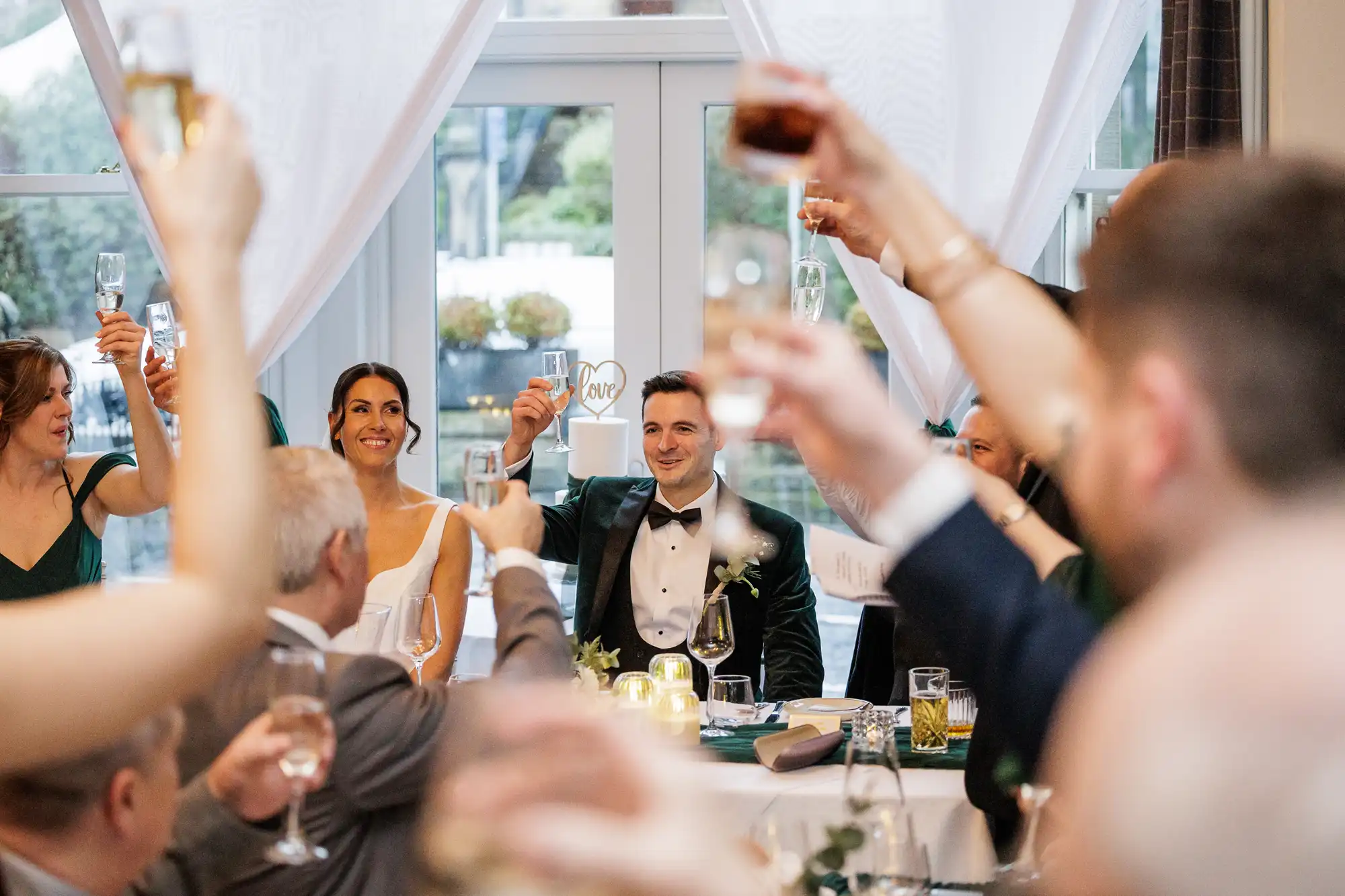 Wedding party toasting with champagne. The groom in a green velvet jacket smiles as hands hold glasses up.
