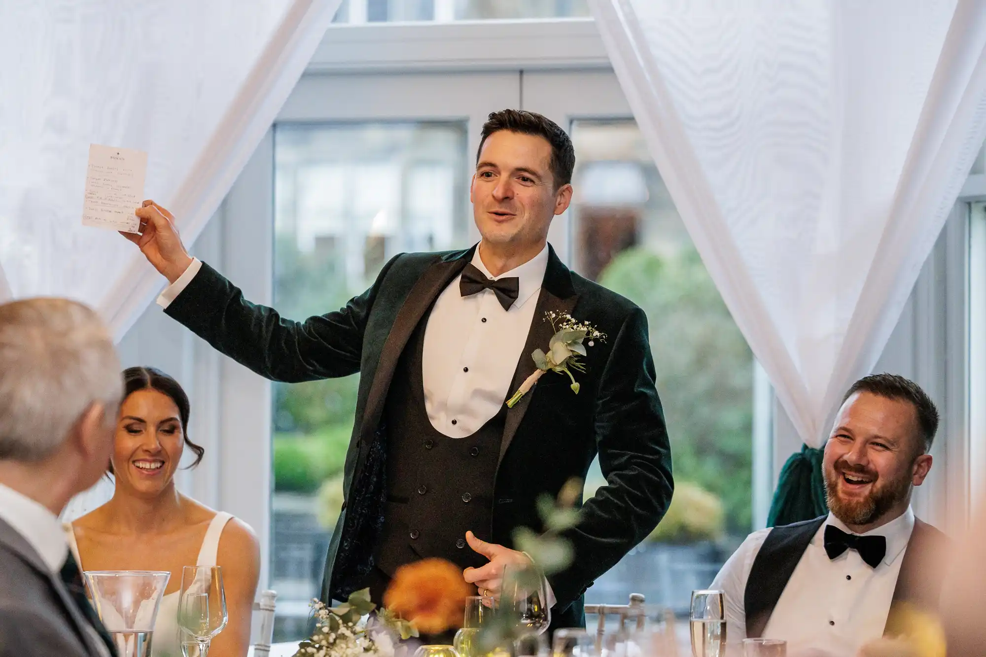 Man in a green velvet tuxedo gives a toast, holding a list. A smiling bride sits at the table, while a laughing man in a vest and bow tie is to his right.