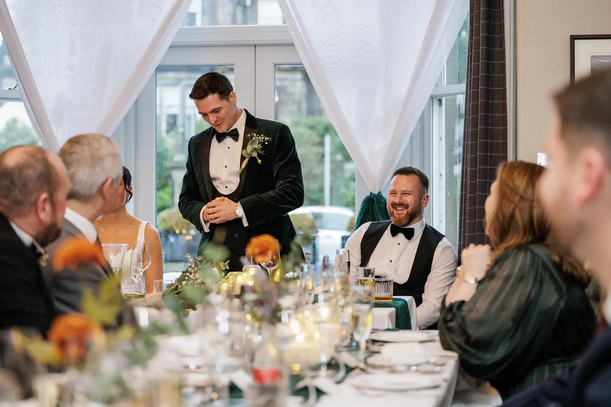 A man in a dark green velvet tuxedo, possibly the groom, stands with his hands clasped, looking down. To his right, a man in a black vest and bow tie laughs heartily. They are seated at a wedding reception table with floral decorations.