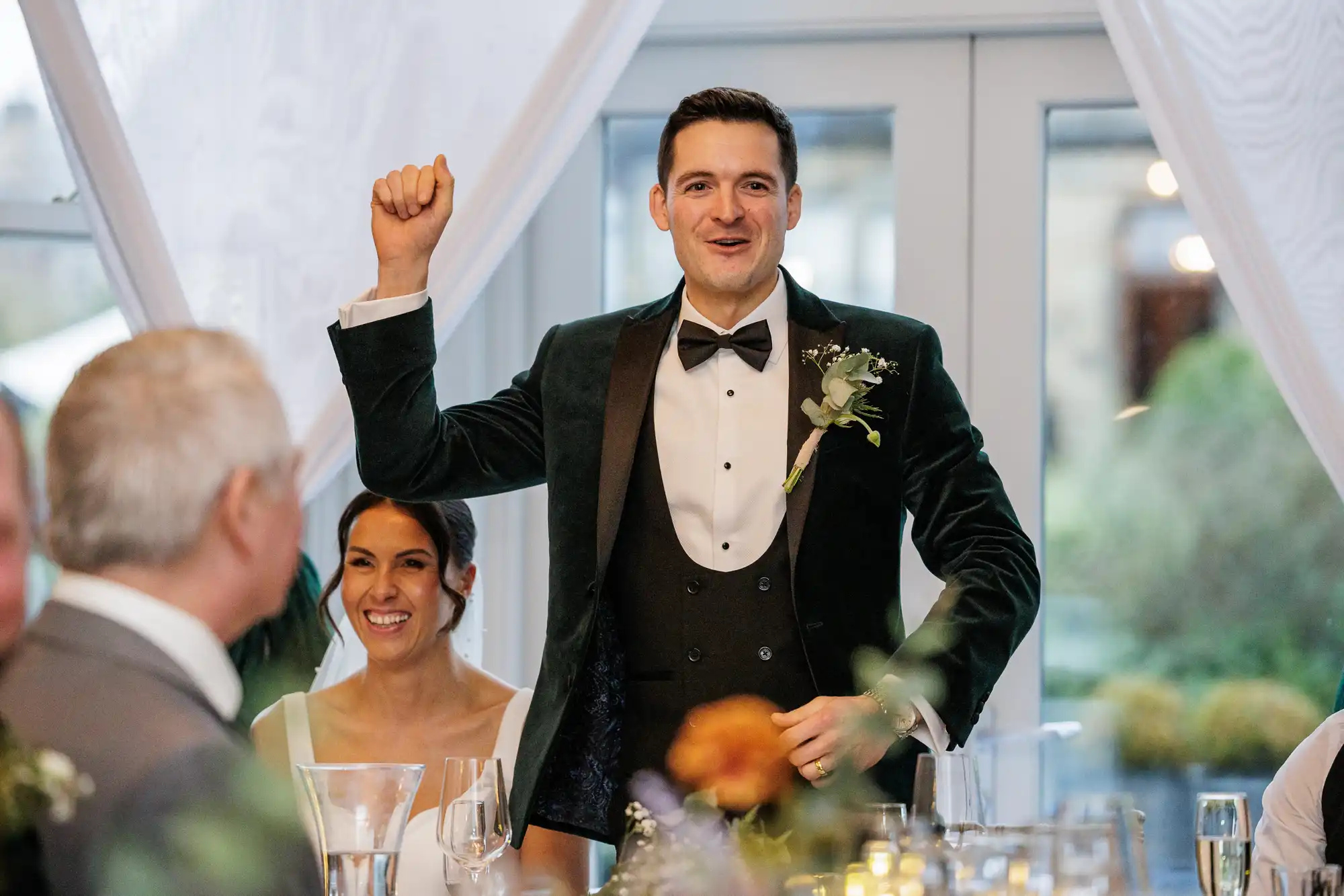 A groom in a velvet green suit and bow tie raises his fist in celebration at a wedding reception, looking joyful. His bride smiles beside him.