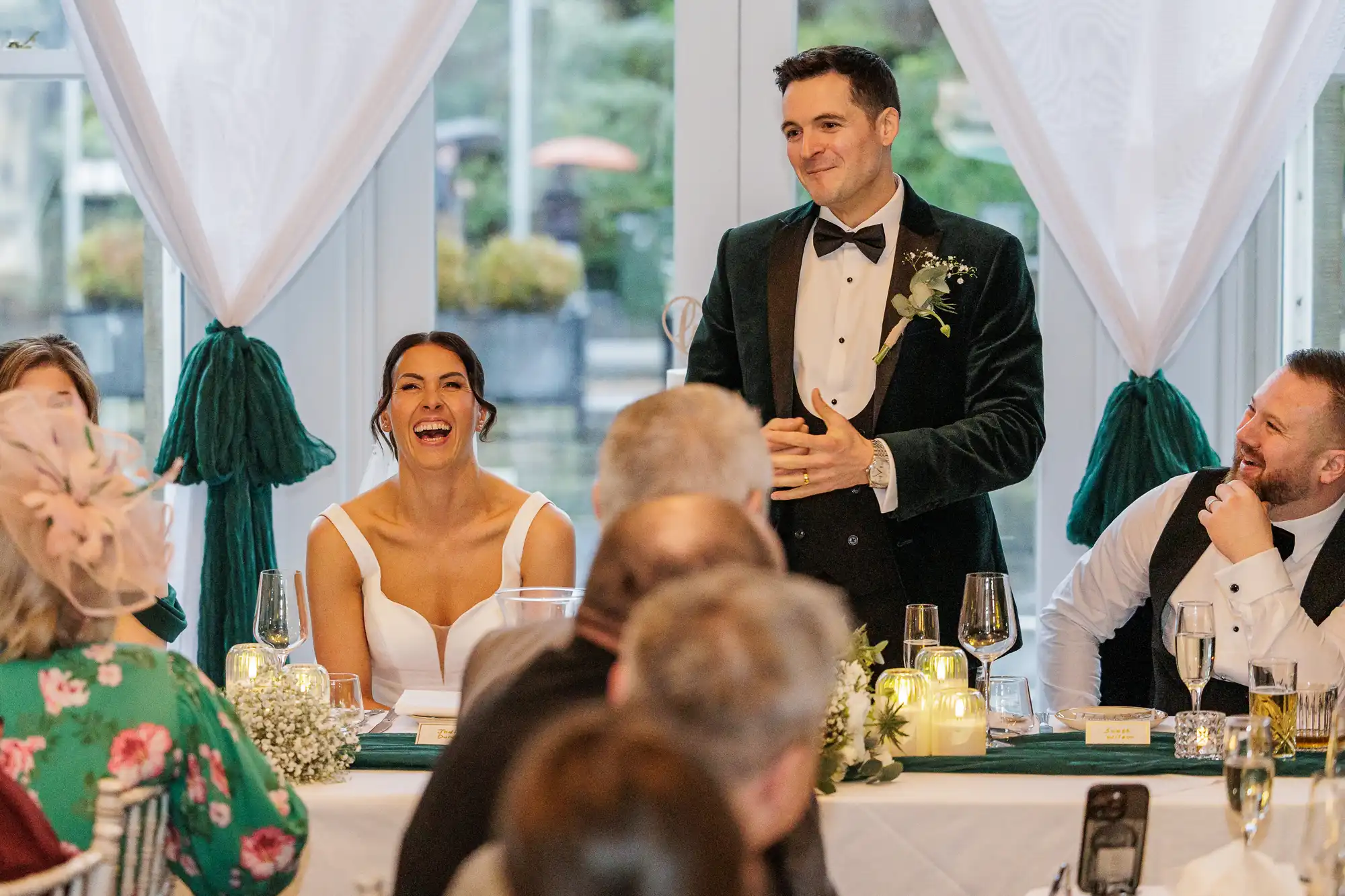 A groom in a velvet tuxedo smiles as he addresses guests at a wedding reception. The bride laughs beside him, wearing a white gown. Guests are seated at a table with floral arrangements and candles.