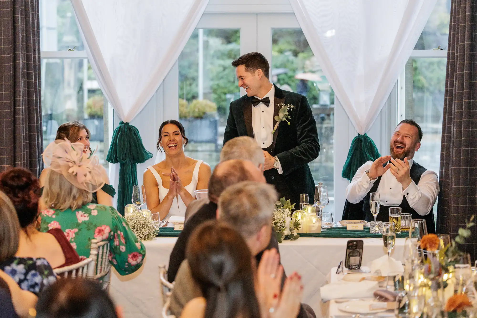 A groom in a velvet jacket and bowtie smiles at his bride, who laughs with her hands clasped. Guests at a wedding reception clap along with the speakers.