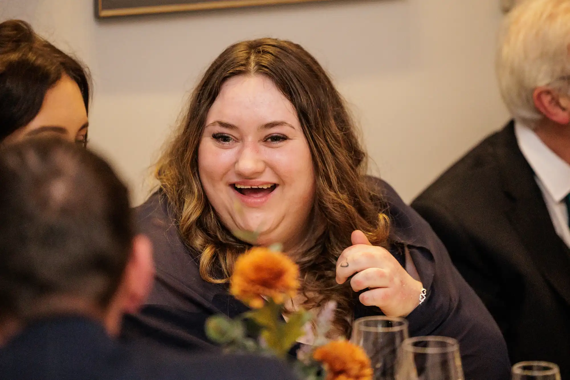 A young woman with long, wavy brown hair laughs heartily at a table. She wears a dark blue top and a silver bracelet.