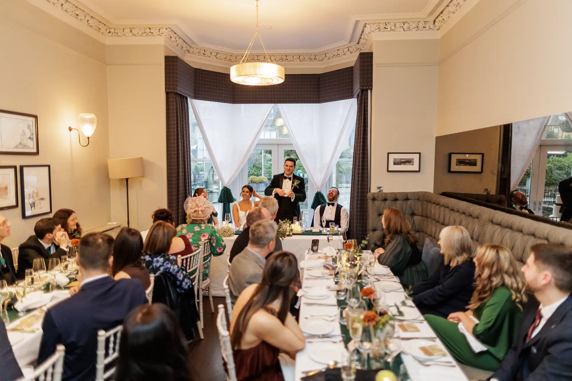 A groom in a tuxedo gives a toast at a wedding reception, while a bride smiles beside him. Guests are seated at tables, with a woman in a green dress and a man in a bow tie laughing.