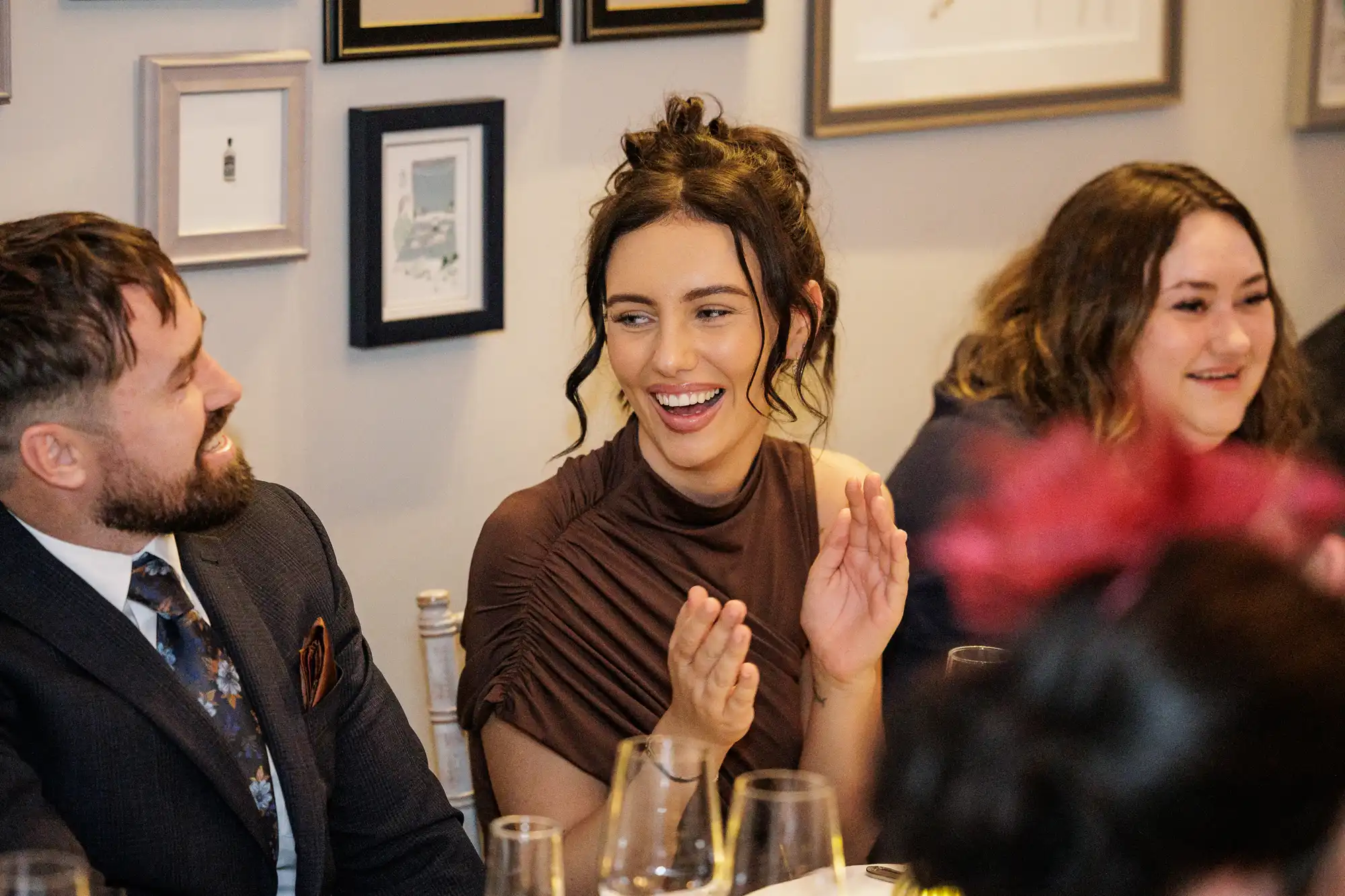 A woman in a brown ruched top smiles and claps at a table. To her left, a man in a suit and patterned tie laughs.